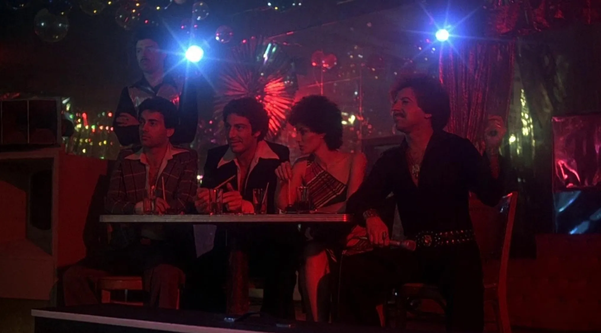 Group of people in 70s movie scenes sitting at a table in a dimly lit club with colorful lights and drinks.