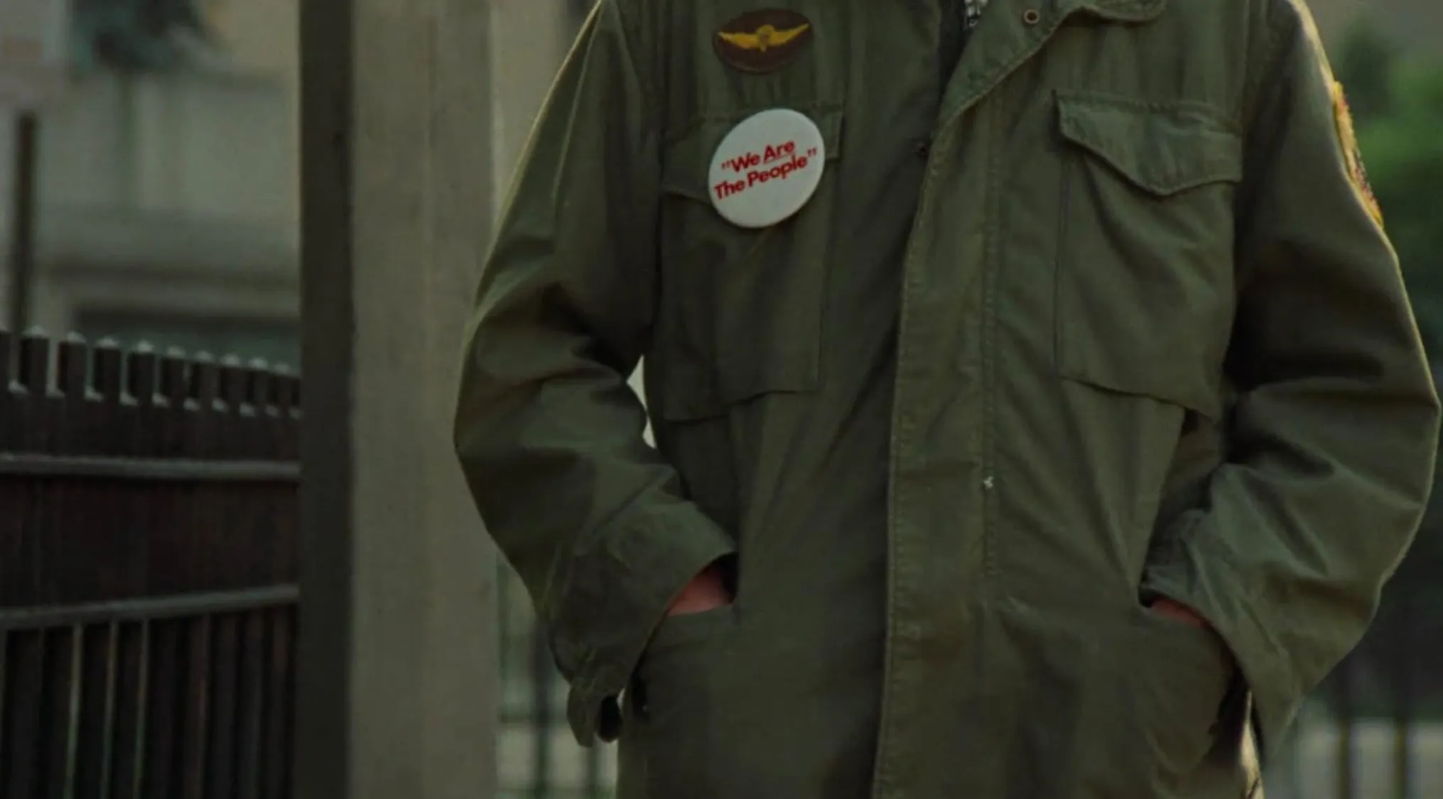 Close-up of a 70s movie scene showing a jacket with a political badge that says We Are The People outdoors.