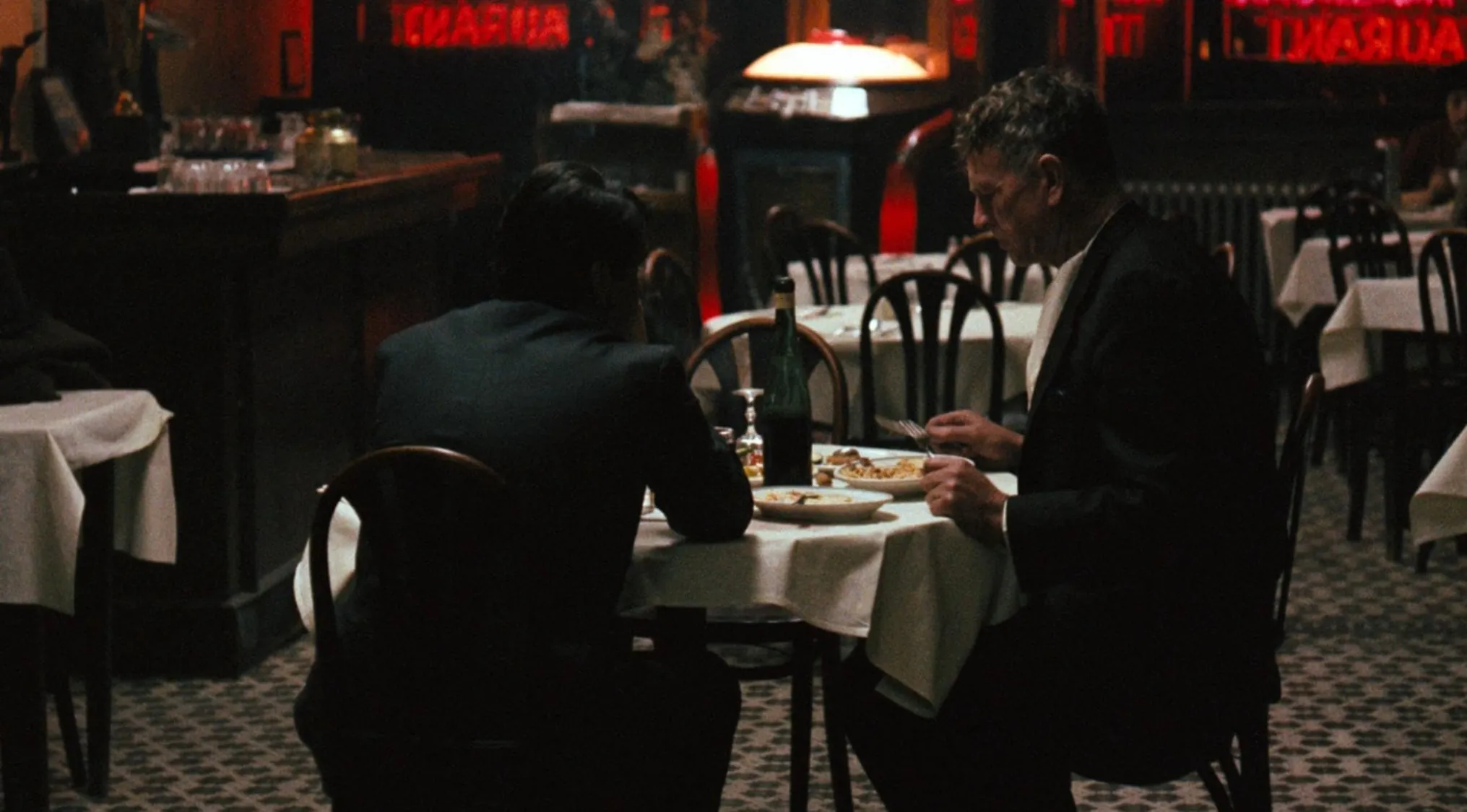 Two men wearing suits dining at a table in a dimly lit restaurant 70s movie scenes from 1970s movies.