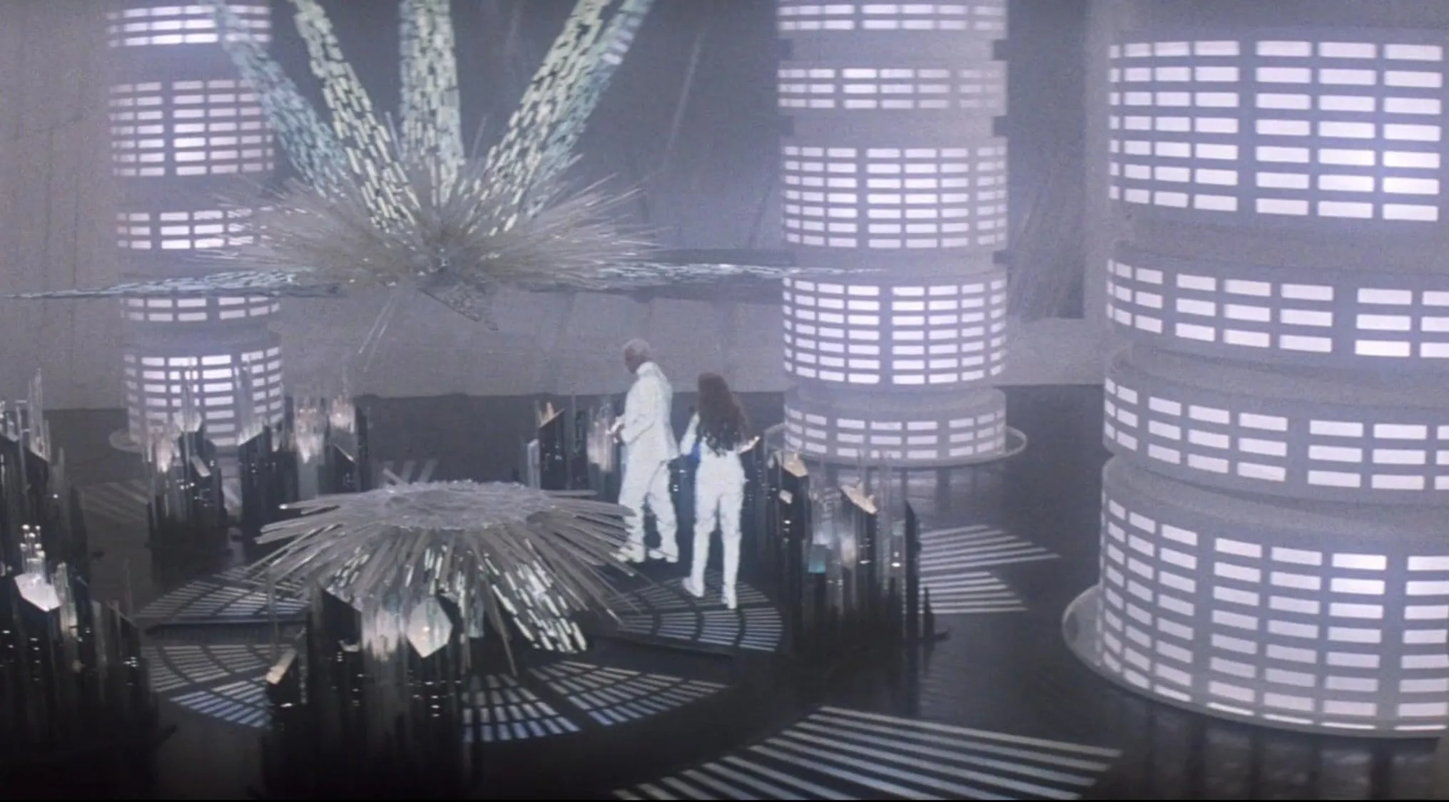 Two characters in white outfits stand amid futuristic columns in a classic 70s movie scene with sci-fi elements.