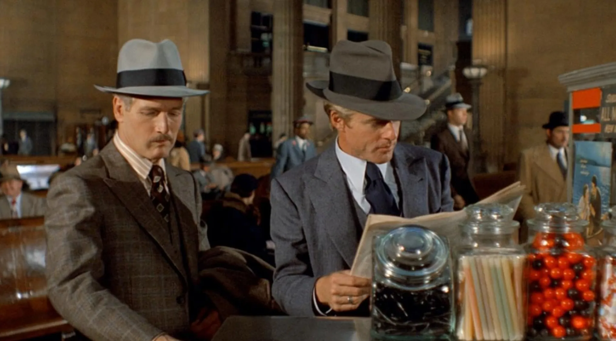 Two men in 70s style suits and hats at a counter with candy jars in a classic 70s movie scene setting.
