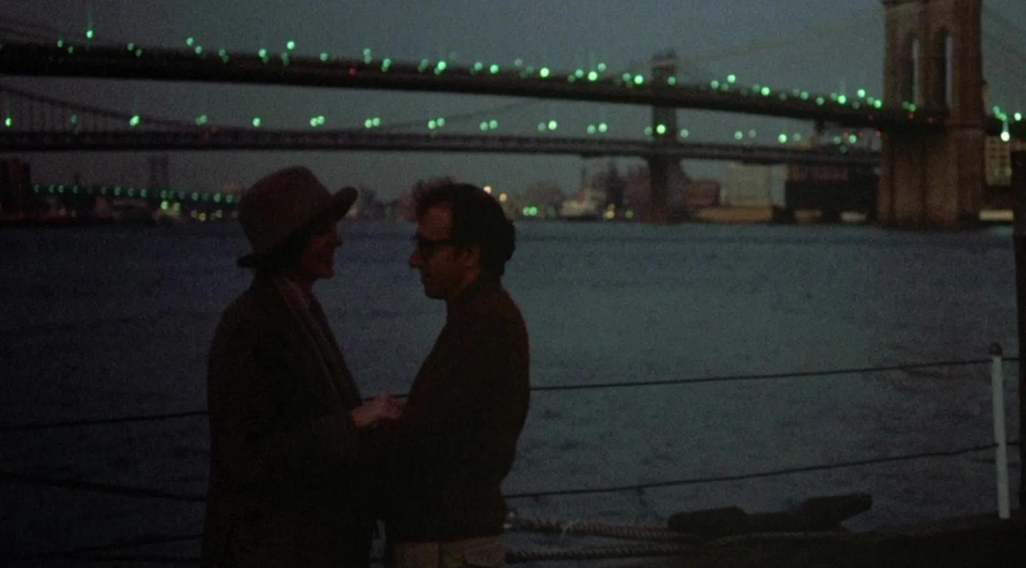 Couple standing by the water at night with a lit bridge in the background in iconic 70s movie scenes.