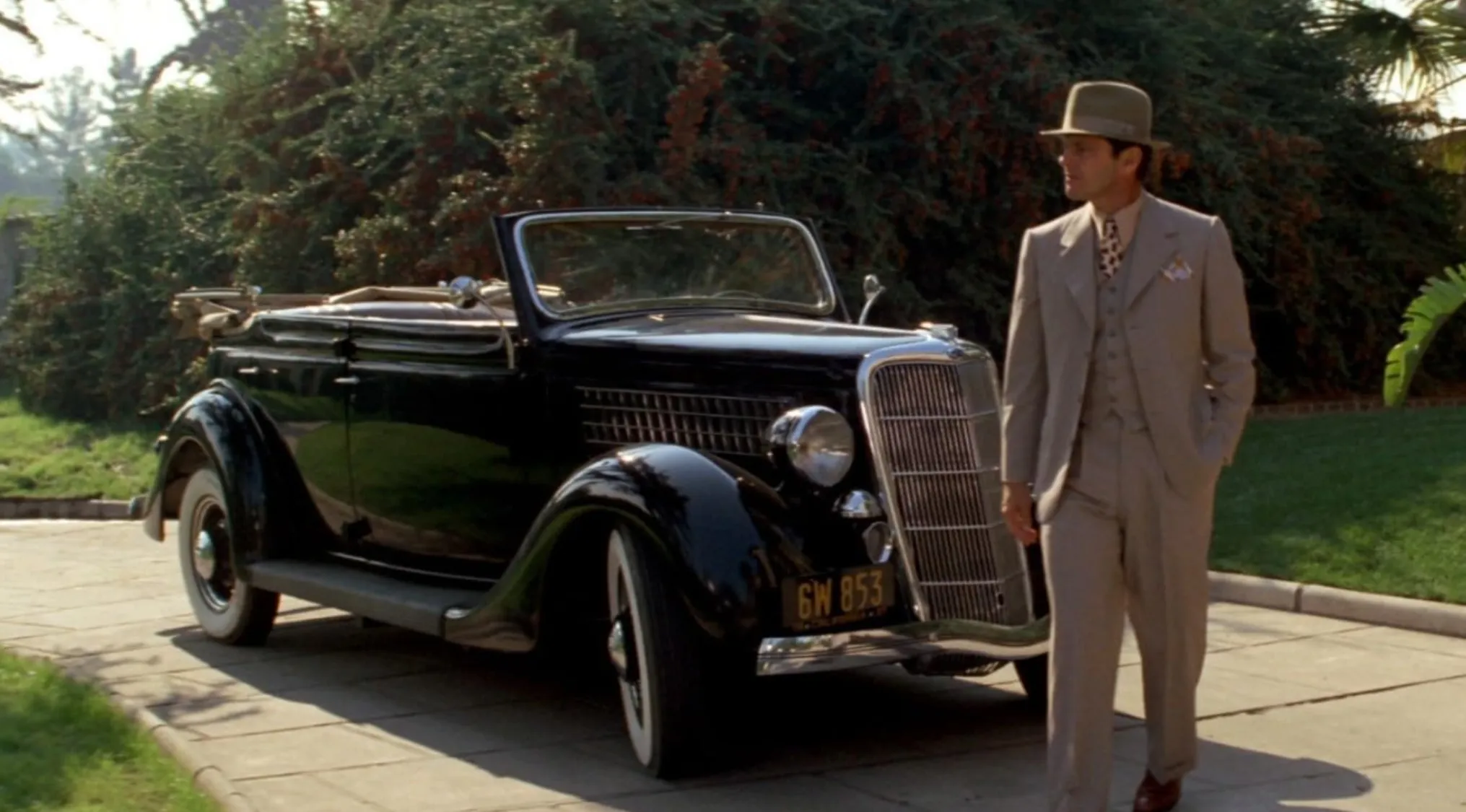 Man in 70s style suit walking next to a vintage black convertible car in a classic 70s movie scene outdoors.