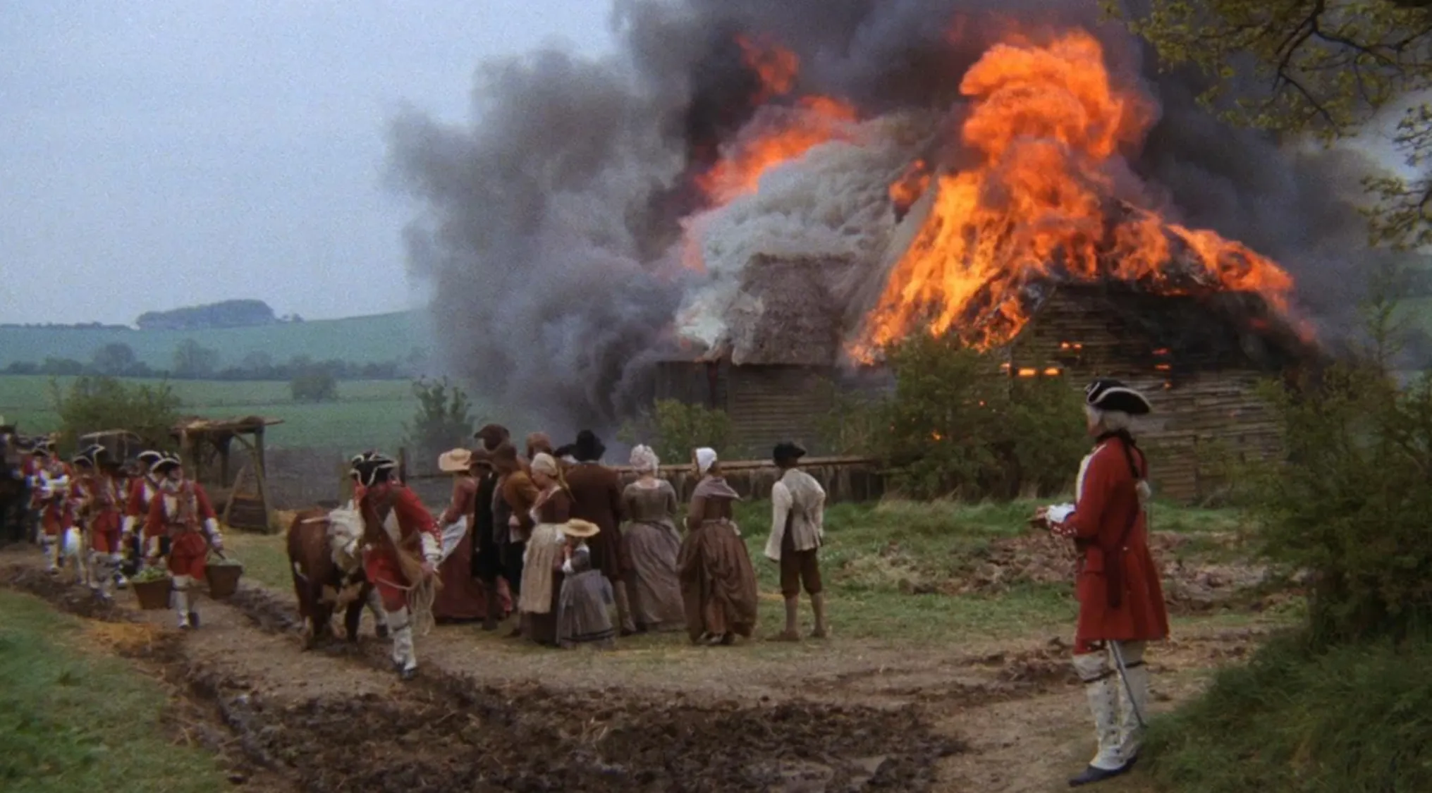 1970s movie scene with villagers and soldiers near a burning thatched cottage in a rural setting.