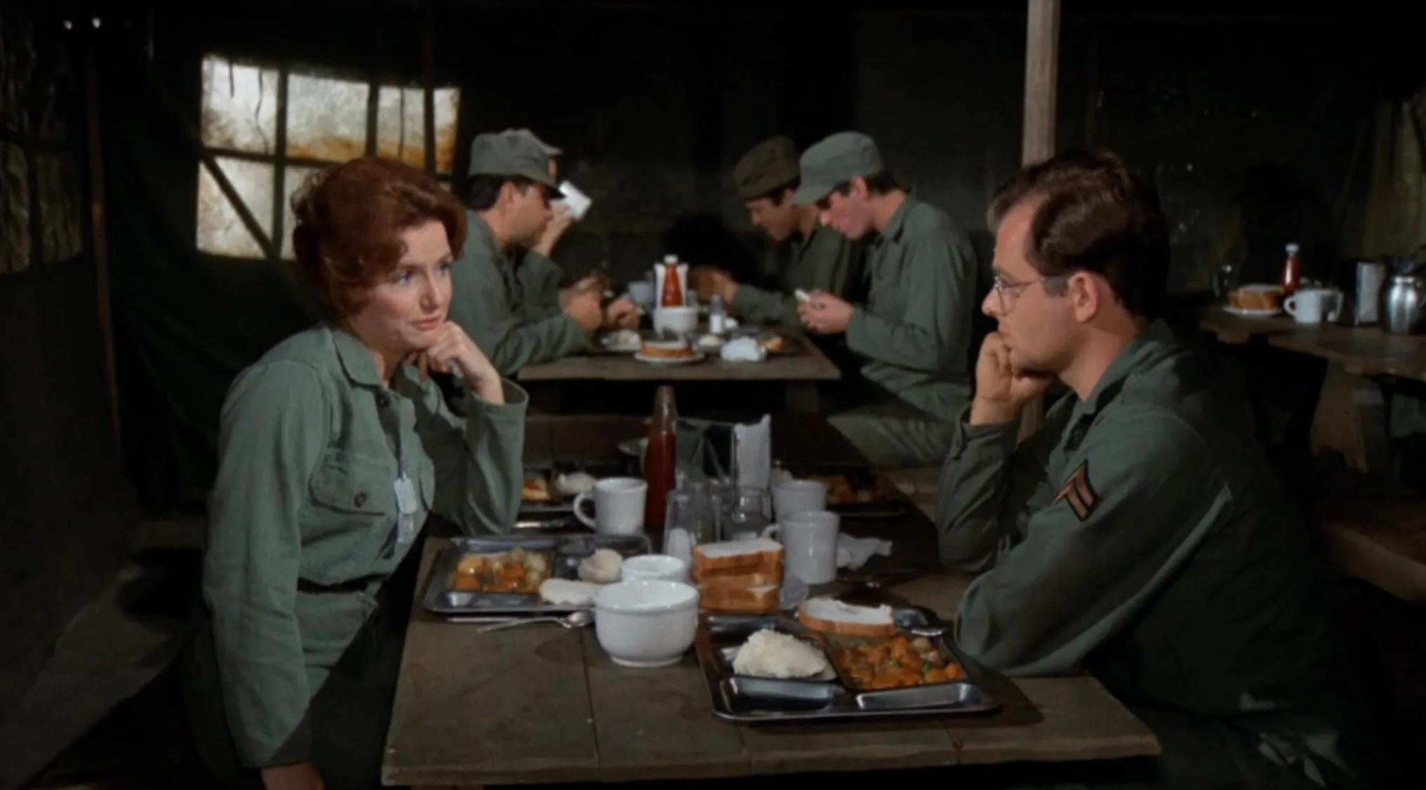 1970s movie scene showing soldiers in uniform having a meal in a makeshift military dining area.