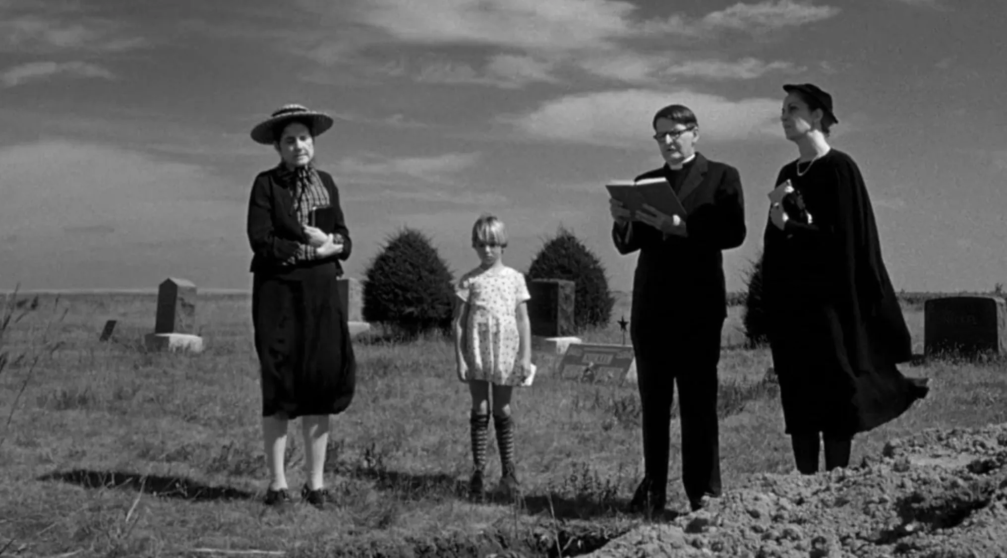 Black and white 70s movie scene with four people standing near a grave in an open field under a cloudy sky.