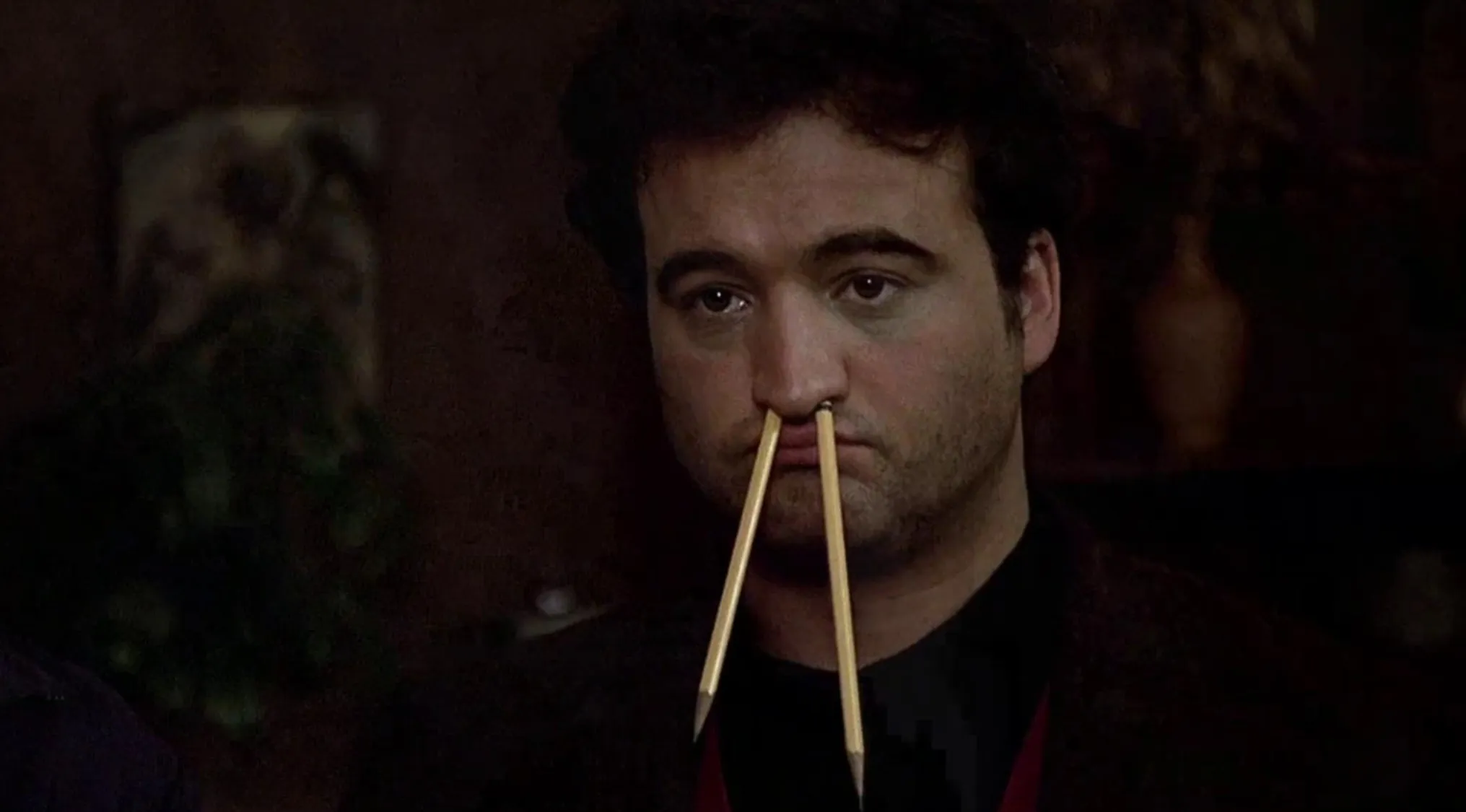 Man with two pencils stuck in his nostrils in a 70s movie scene guessing game setting.