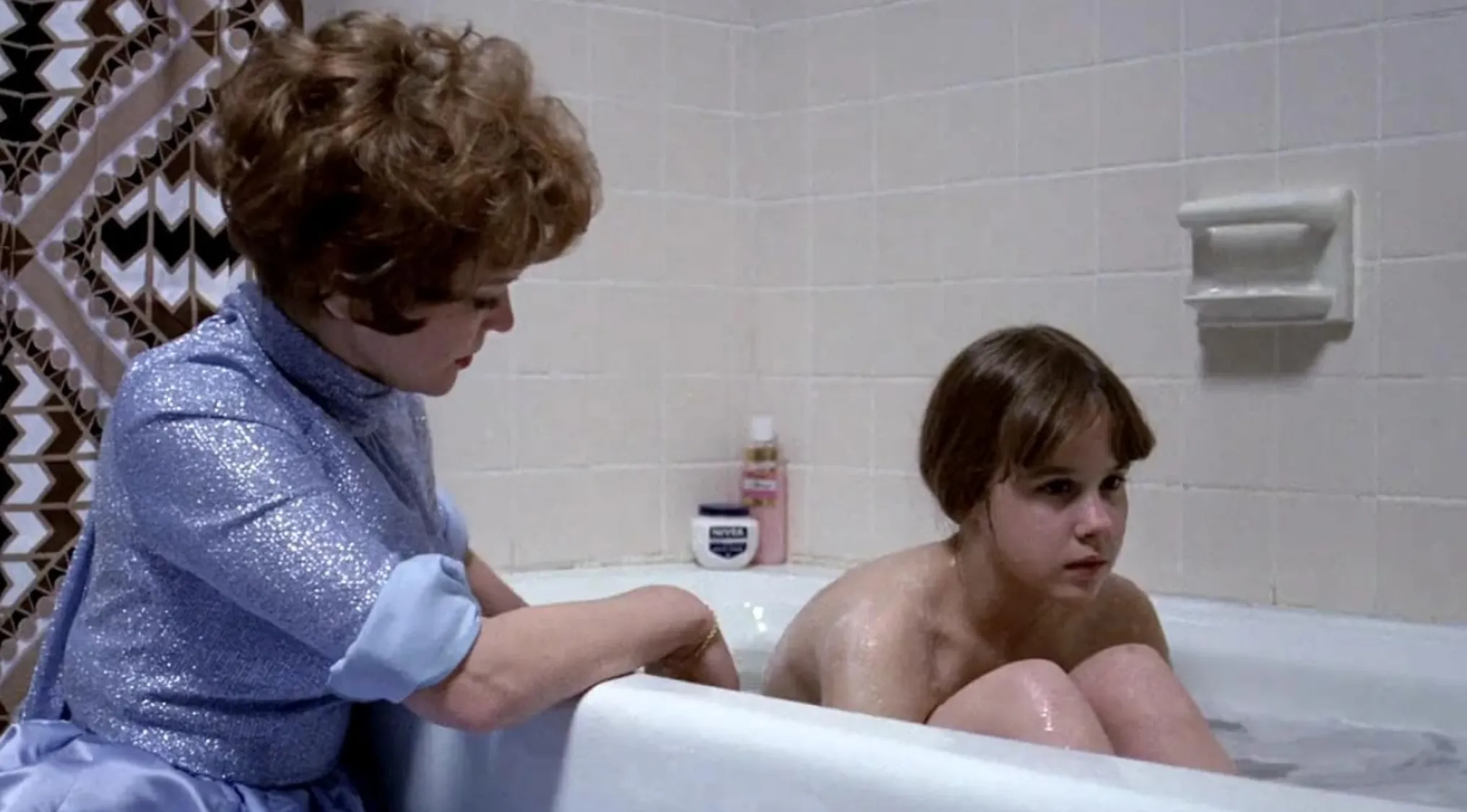 Woman in a blue blouse tending to a young girl in a bathtub in a 70s movie scene with iconic mood and setting.