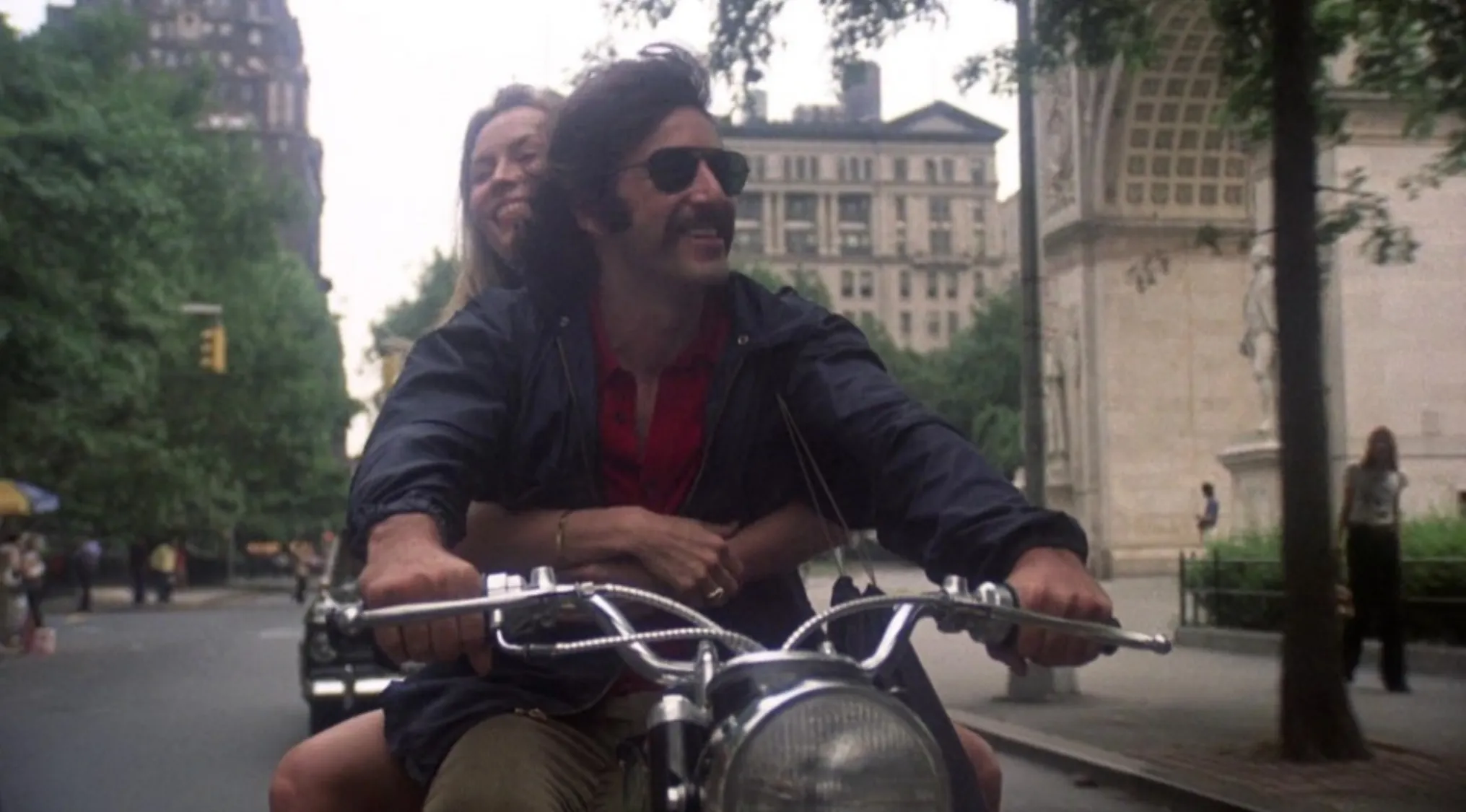 Couple riding a motorcycle in a popular 70s movie scene, set in an urban street with buildings and trees.