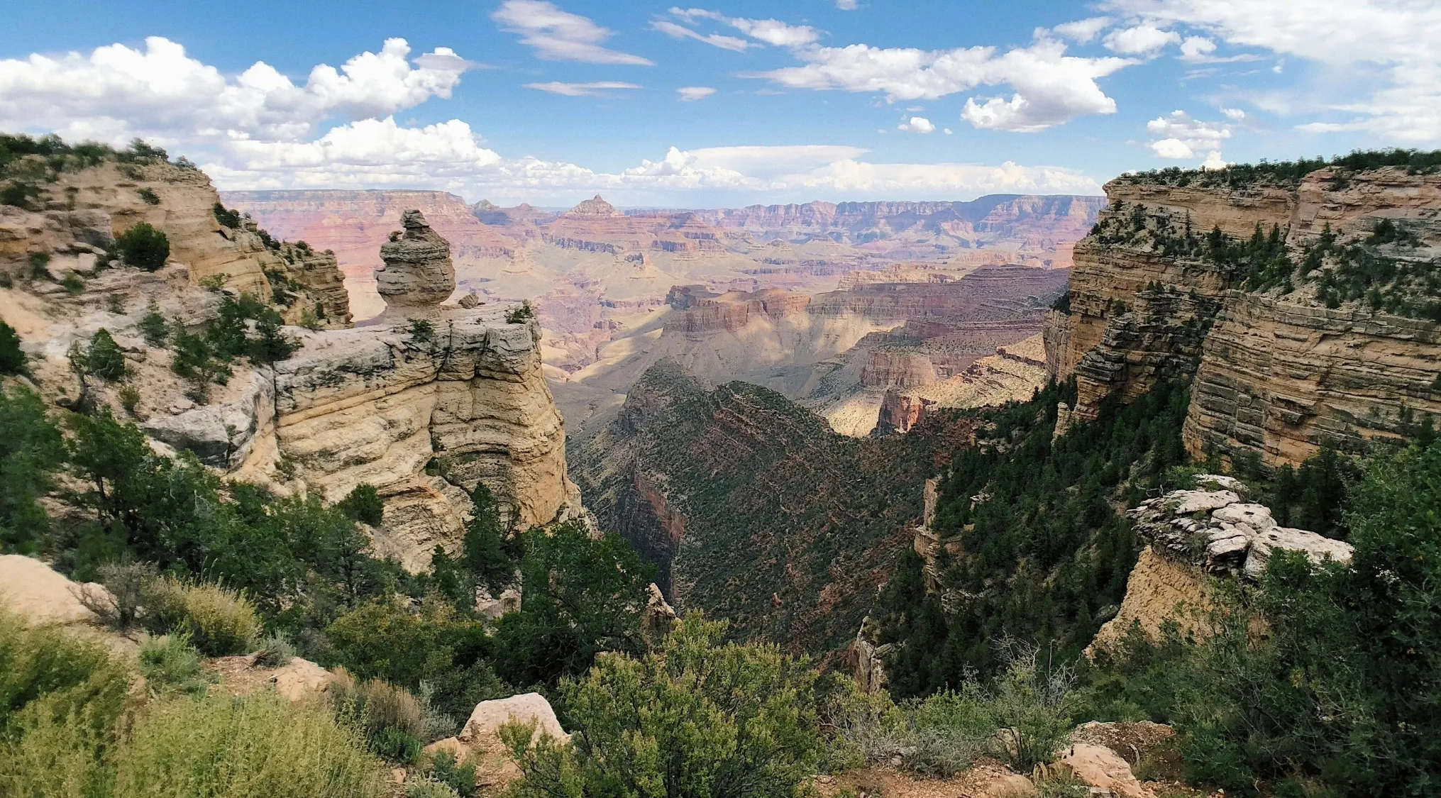 Grand Canyon landscape view with layered rock formations, deep valleys, and scattered greenery under a partly cloudy sky.
