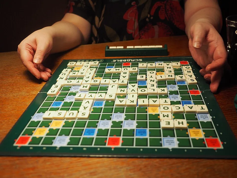 Hands arranging tiles on a Scrabble board during a word game played on a wooden table.