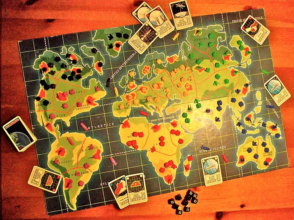 Risk board game map with colored player pieces and strategy cards on a wooden table during gameplay.