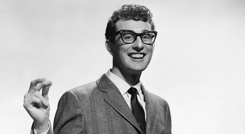 Black and white photo of Buddy Holly smiling, wearing glasses, a suit, and tie, known for rock and roll music.