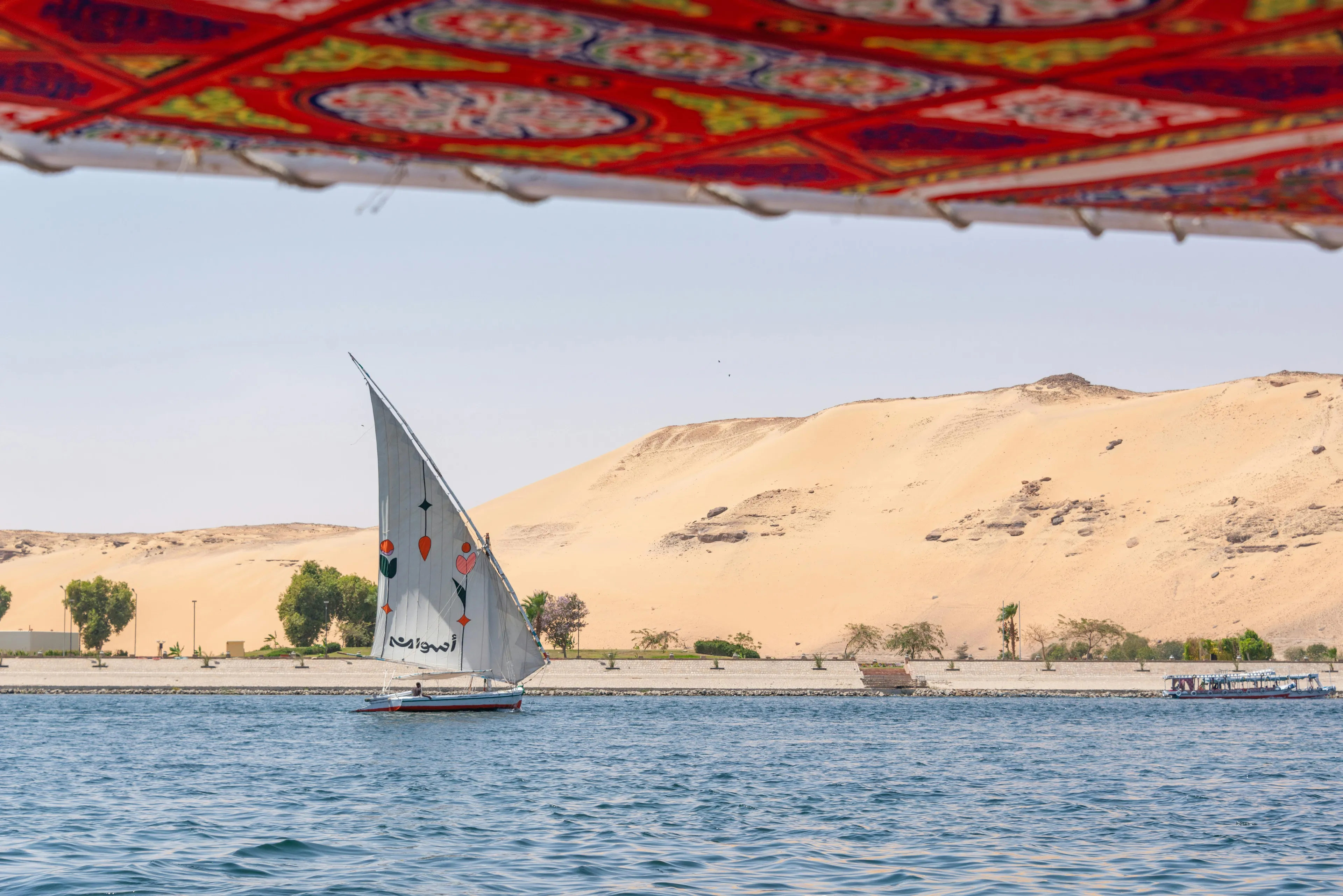 Sailboat on the Nile river flowing through Cairo with desert hills and trees along the riverbank under a colorful canopy.