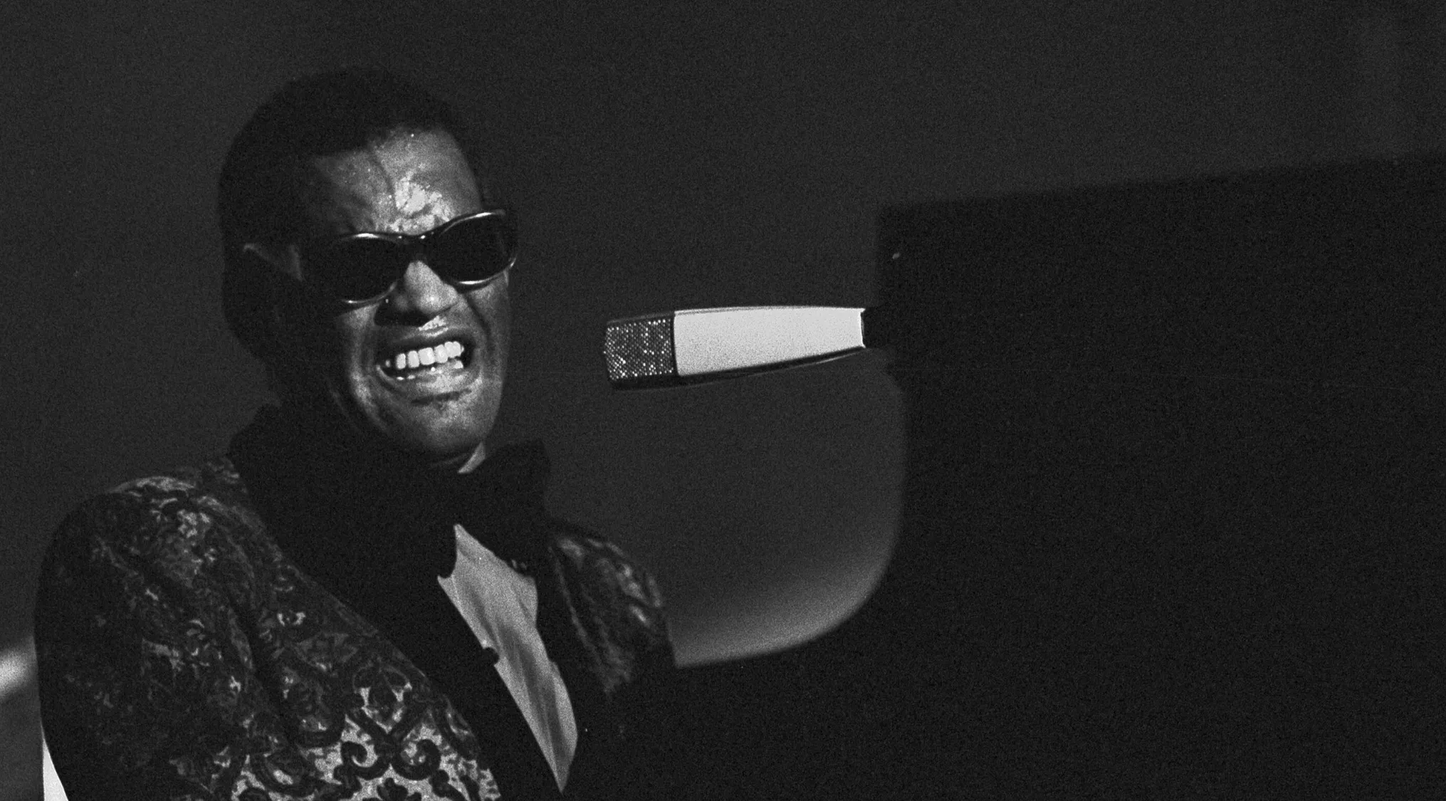 Ray Charles playing piano on stage, wearing sunglasses and a patterned jacket during a live performance.