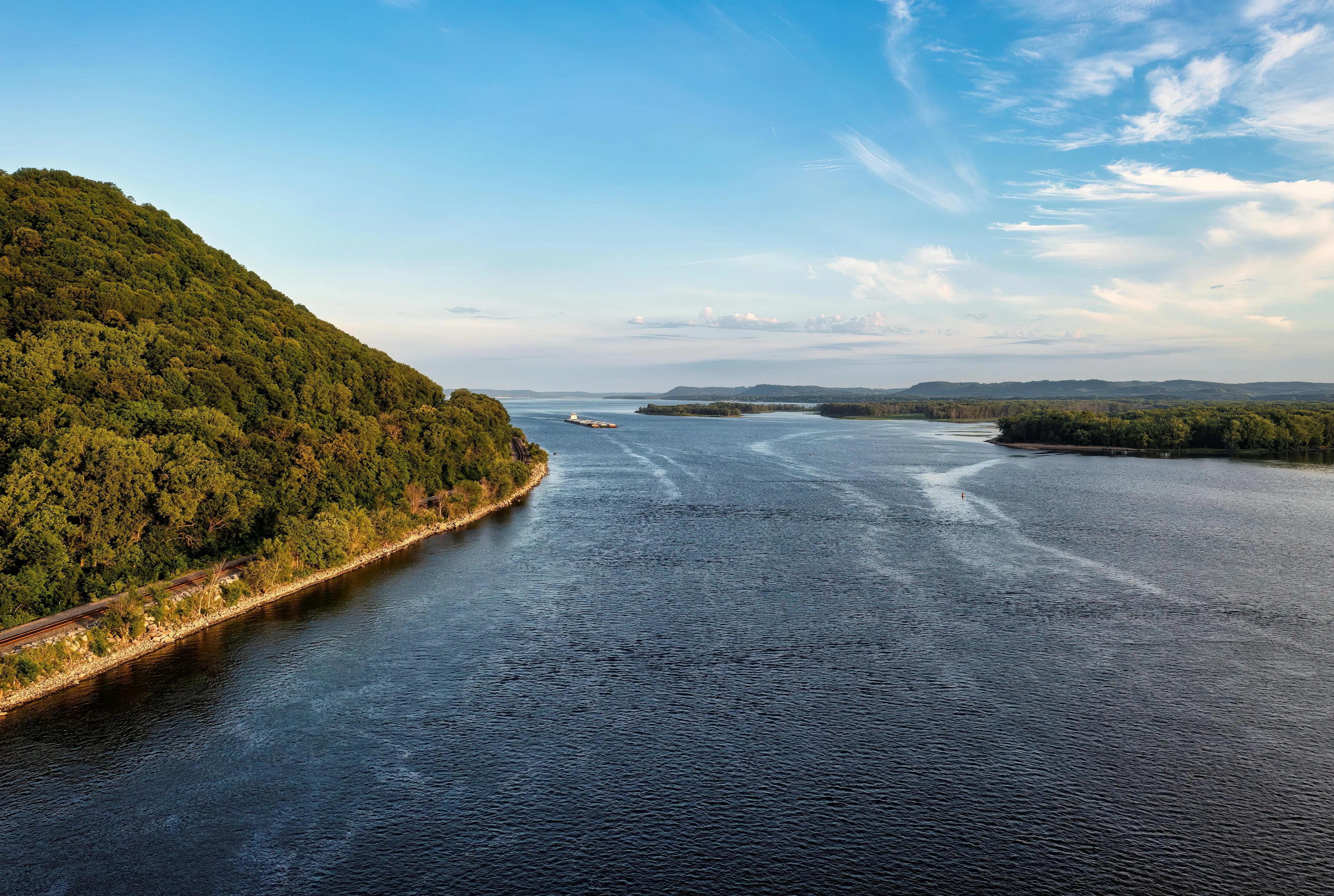 Wide view of a US river nicknamed Ol' Man River, The Big Muddy, and Old Blue flowing past green forested hills under a blue sky.