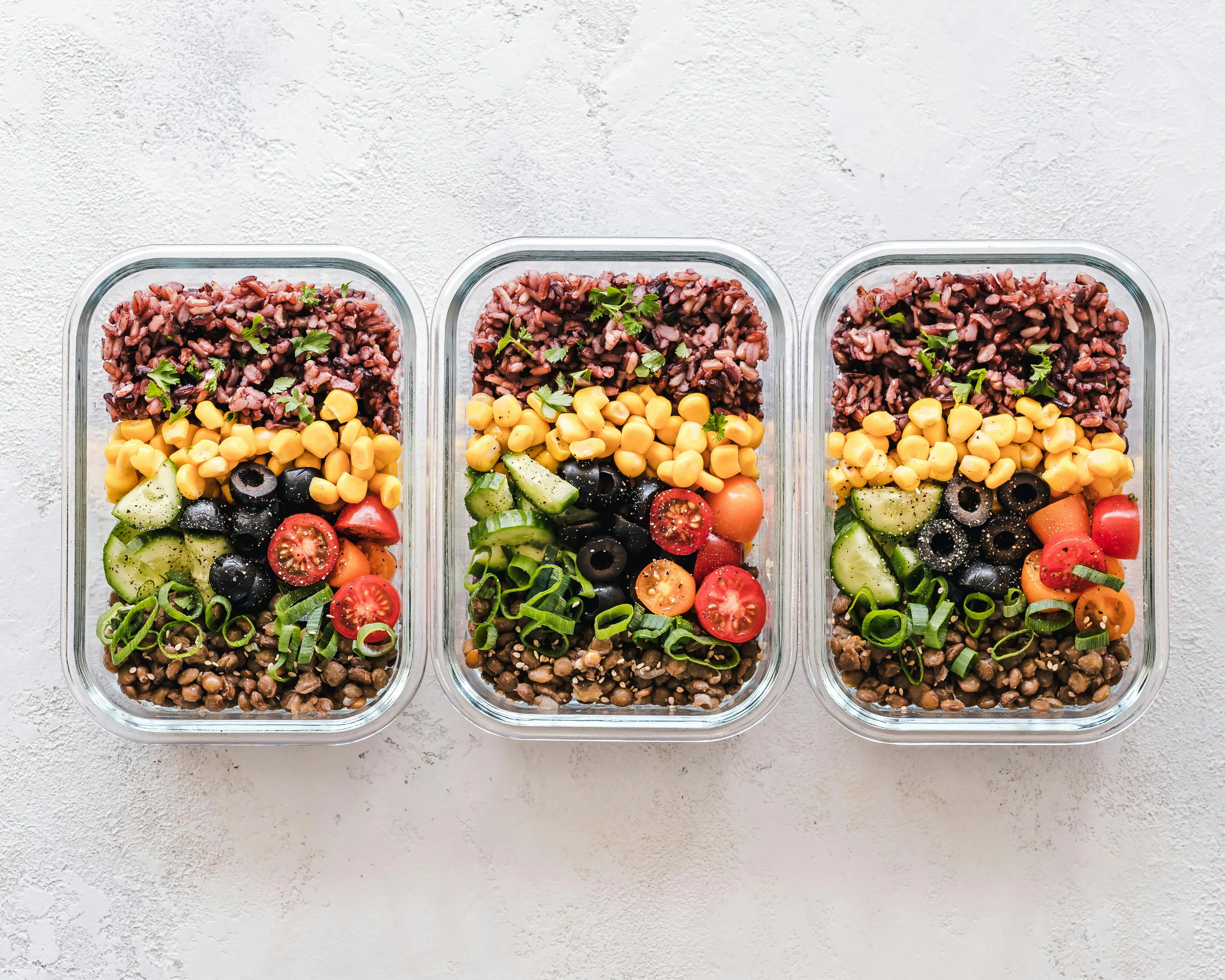 Three meal prep containers with healthy ingredients including corn, olives, cherry tomatoes, cucumbers, lentils, and rice for macronutrient meals.