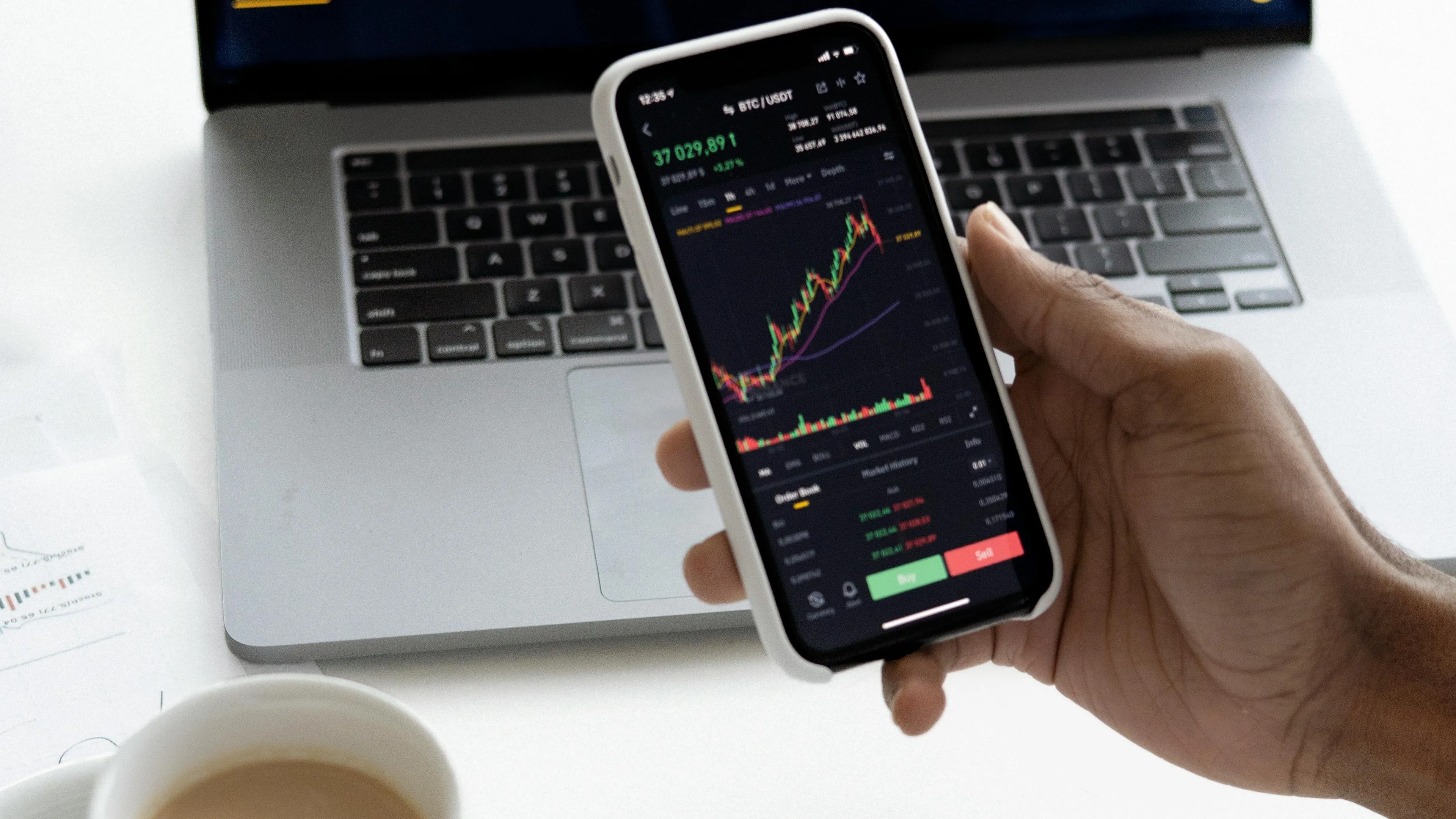 Hand holding smartphone showing stock and bond market charts with laptop and coffee cup in the background.