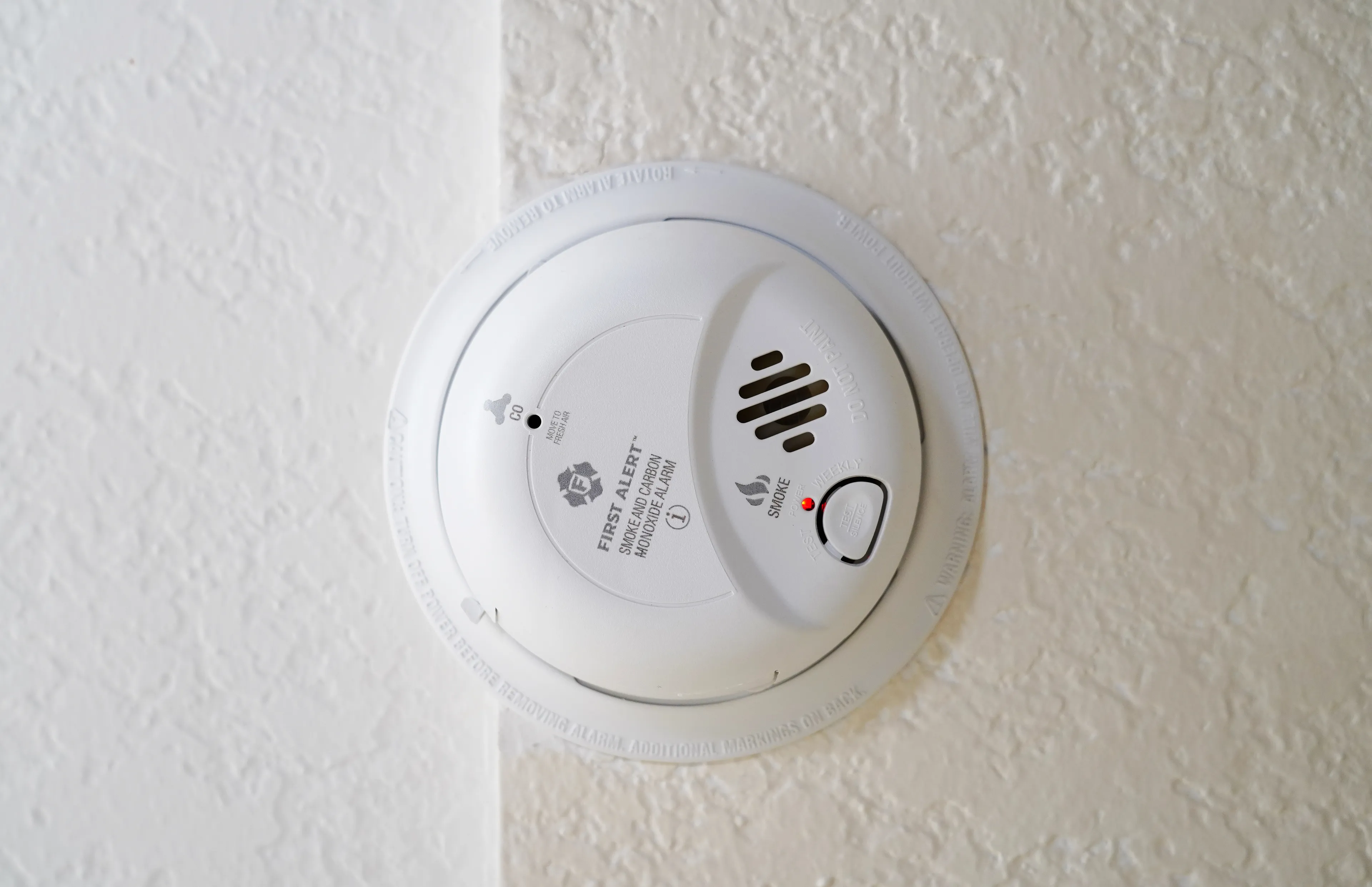 Smoke detector mounted on ceiling with test button and red indicator light on textured white wall.