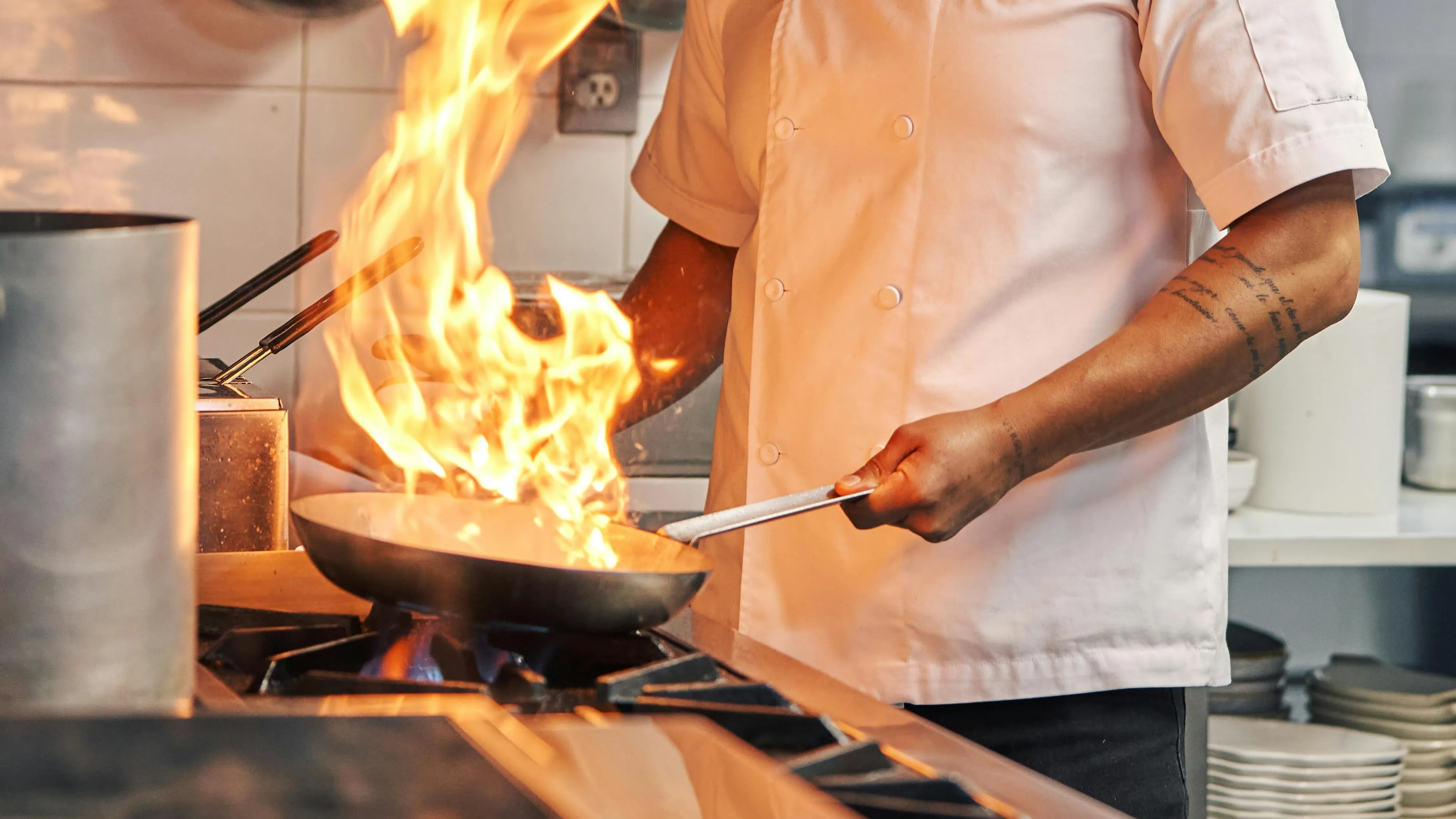 Chef in a white coat cooking with grease fire flame rising from a pan on a kitchen stove showing fire safety risks.