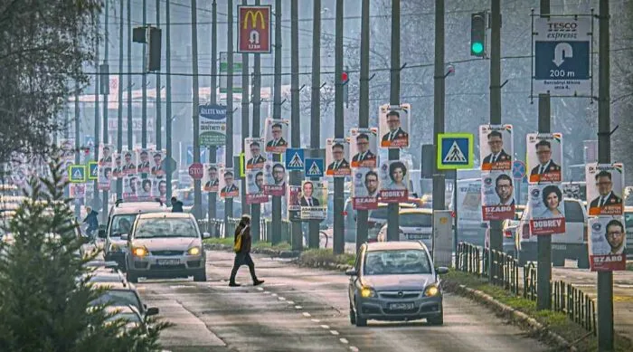 Urban street filled with election posters on poles, cars driving and a pedestrian crossing in dystopian urban hell scene.