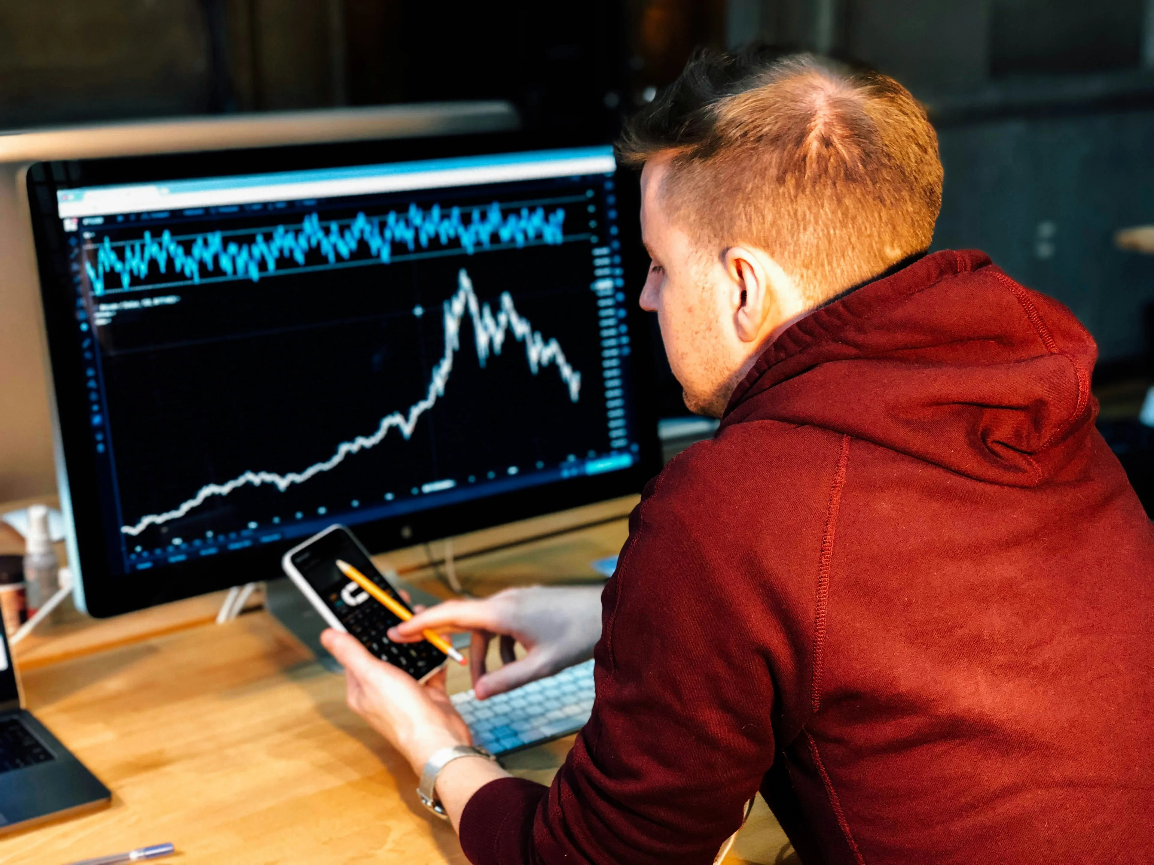 Man in a red hoodie analyzing GDP data on computer screen and calculator in a focused general knowledge quiz setting.