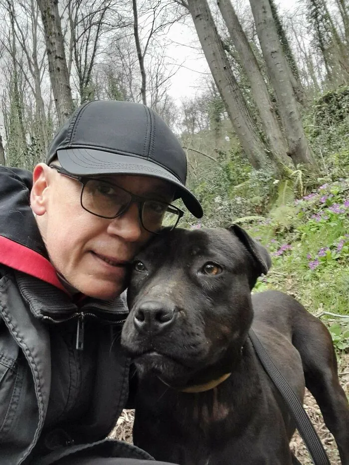 Man in glasses and black cap posing closely with a black rescue dog during a forest walk, symbolizing forever homes for dogs.