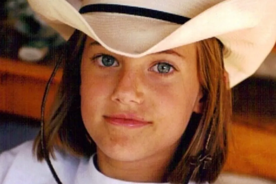 Young girl wearing a white cowboy hat and white shirt, looking directly at the camera in a casual setting.