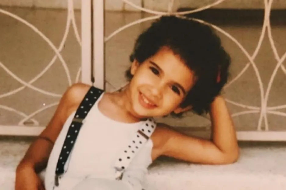 Young girl smiling and reclining with arm behind head, wearing white top and black polka dot suspenders in casual setting.