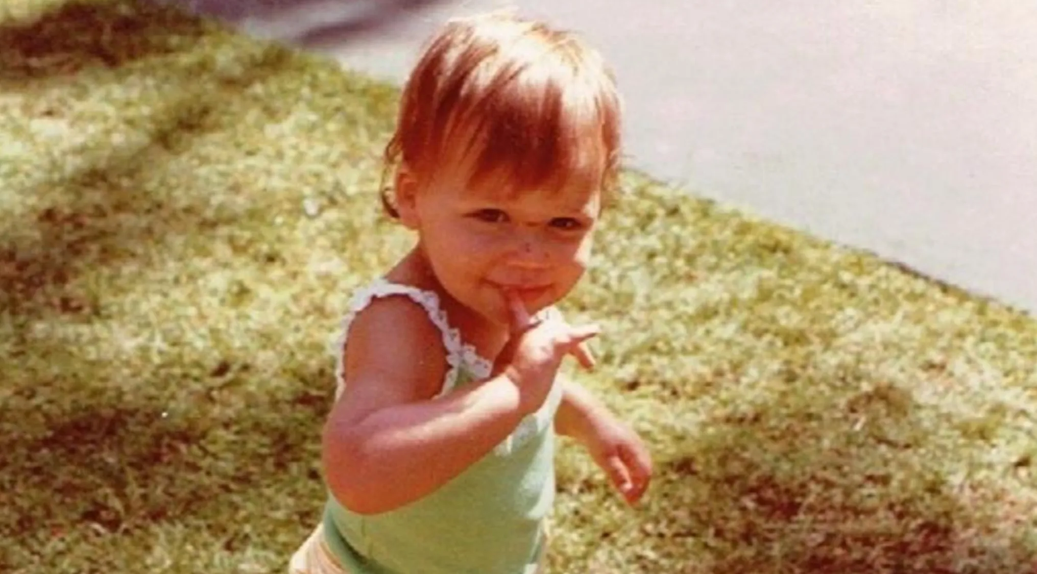 Young child outdoors on grass, smiling with finger near mouth, part of celebrities as kids photo series.