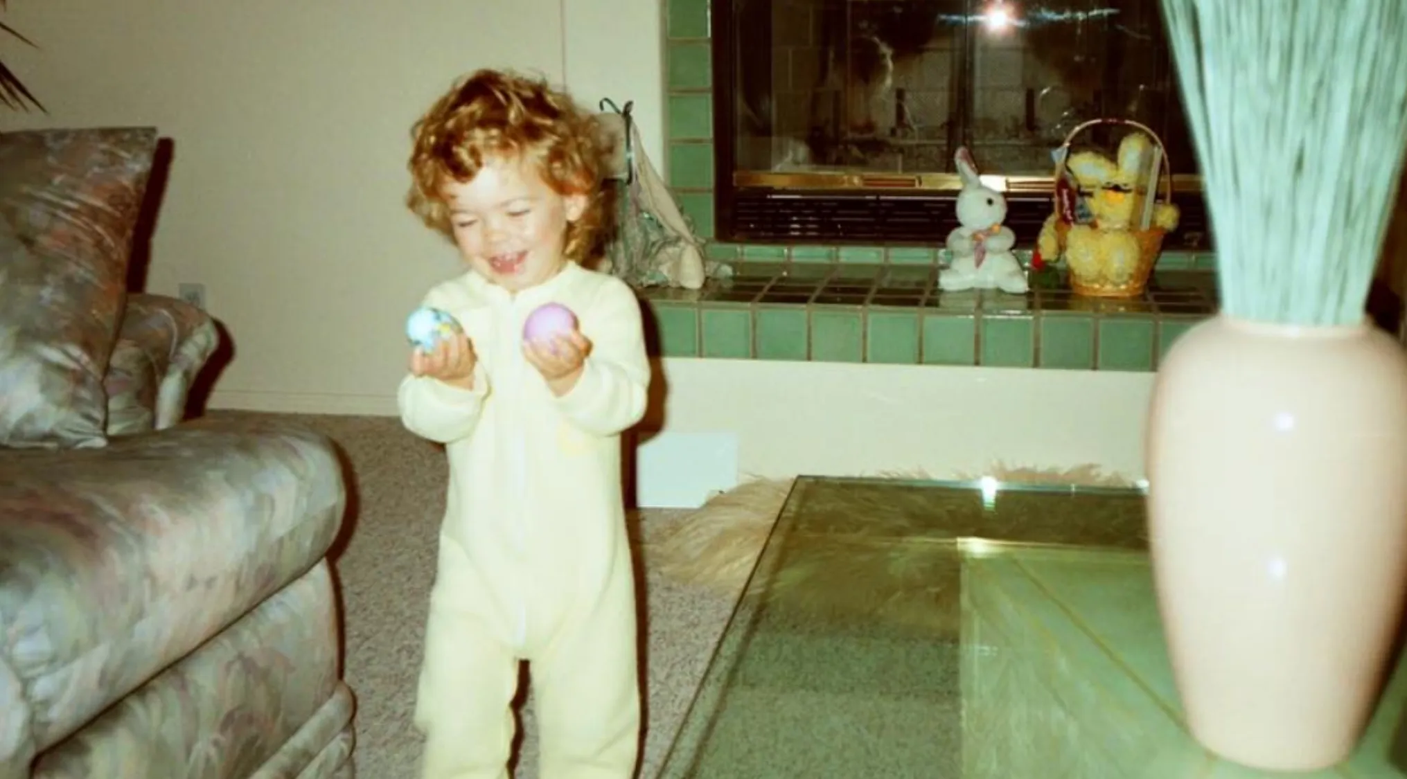 Young child celebrating Easter indoors holding colorful eggs with fireplace and Easter decorations in background