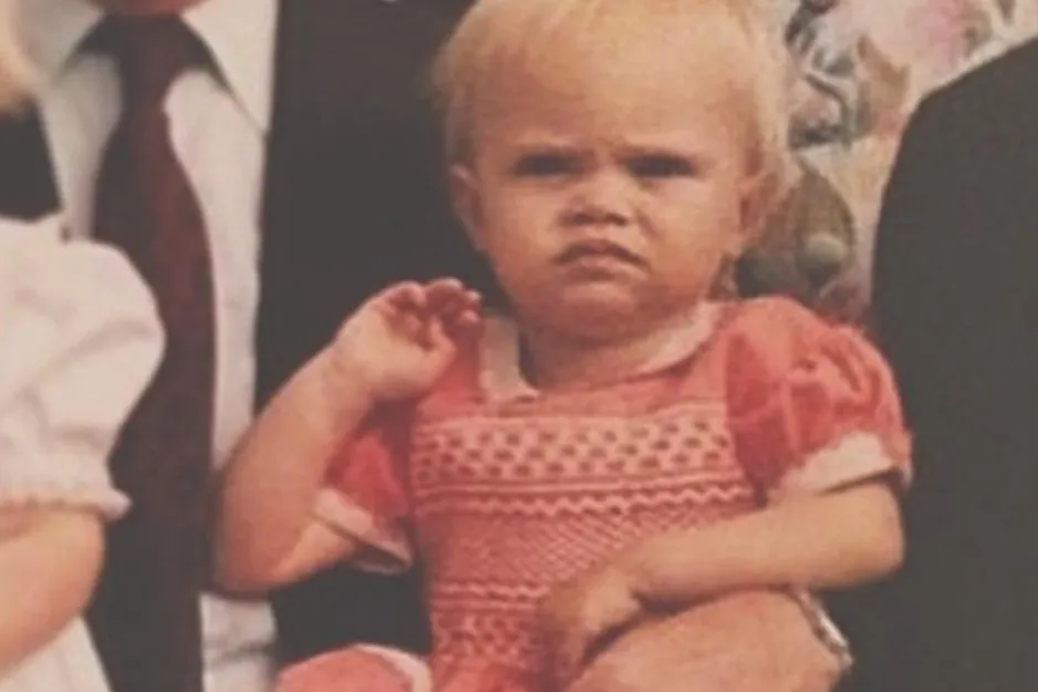 Toddler with a serious expression dressed in a red patterned dress, posing among adults in formal attire.
