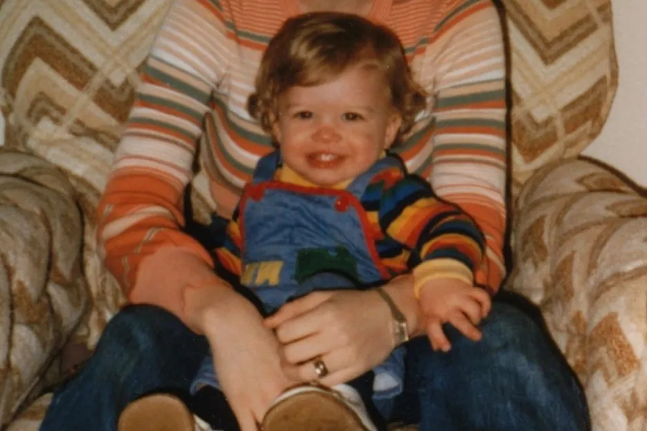Toddler sitting on a striped sofa wearing a colorful outfit, smiling and being held by a person in a striped long sleeve shirt.