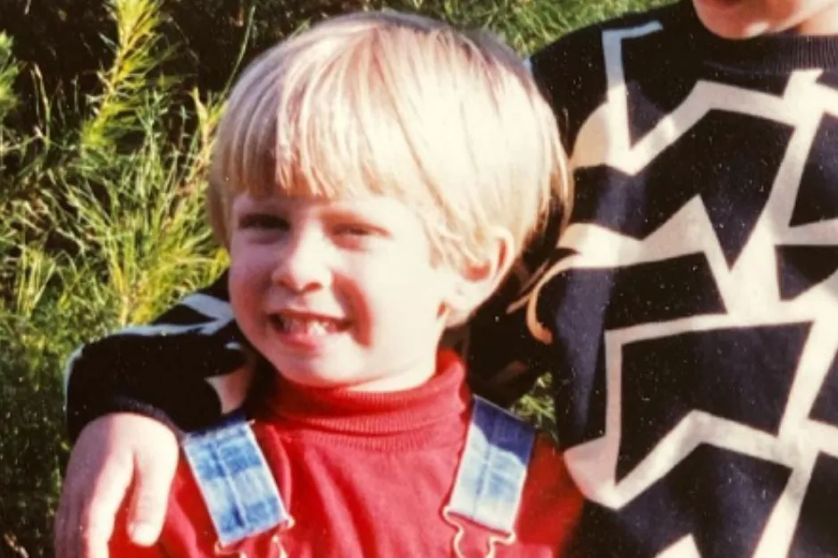 Young boy with blonde hair wearing red shirt and denim overalls outdoors, try to spot Justin Timberlake in the photo