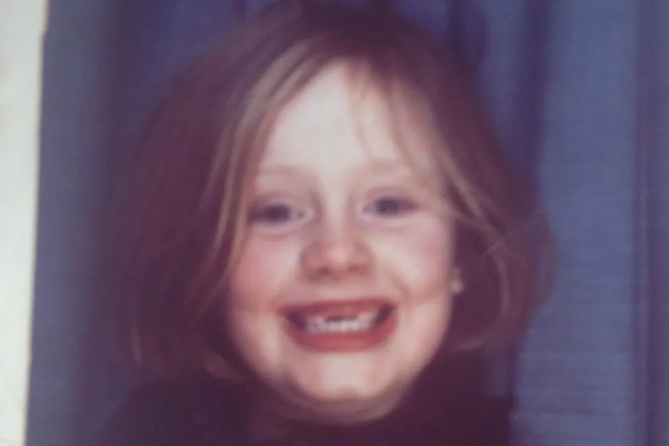 Adele smiling in a casual outfit, standing among a group of people, highlighting which one is Adele in the image.