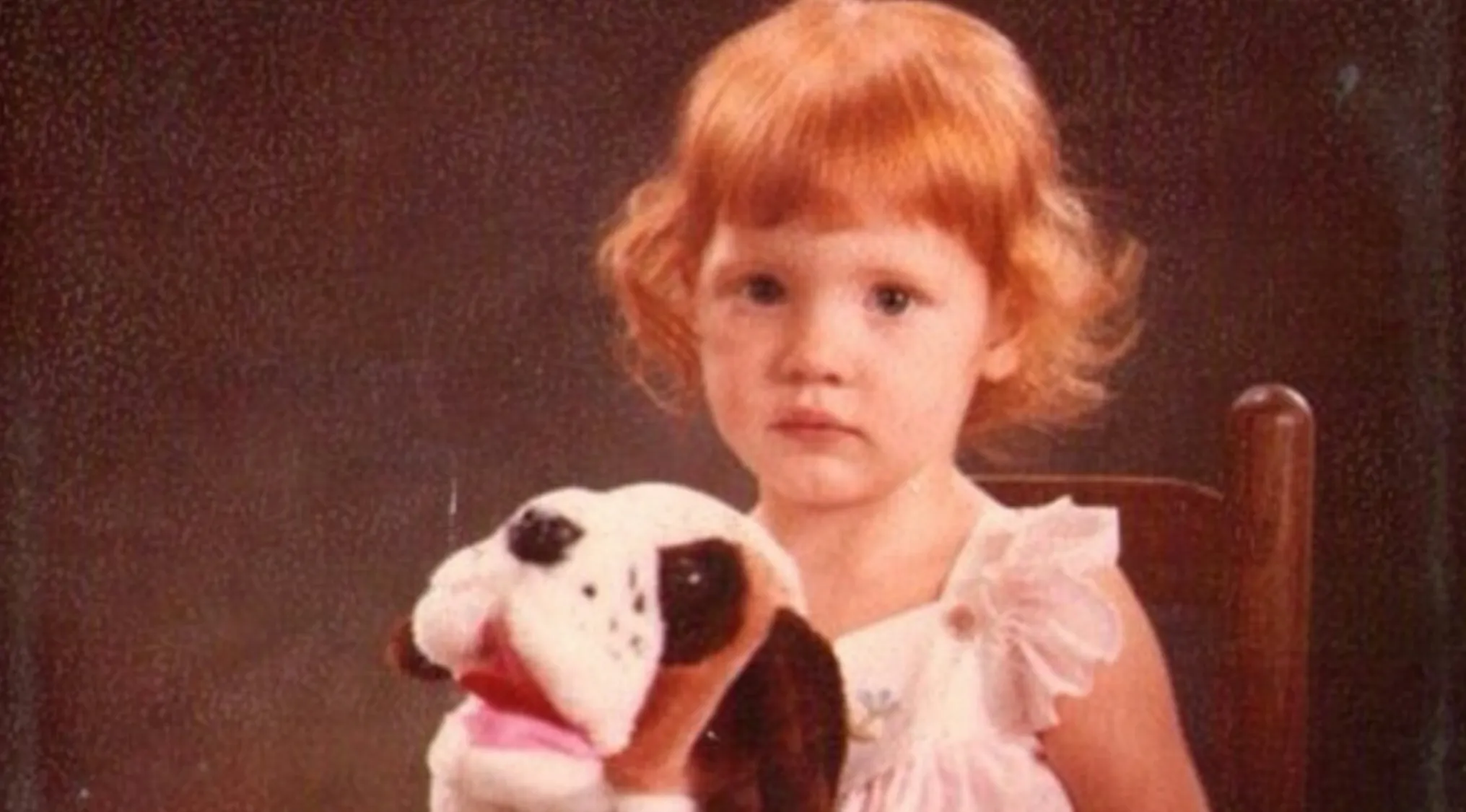 Red-haired child holding a stuffed dog toy sitting on a chair in a vintage style photo from celebrities as kids.
