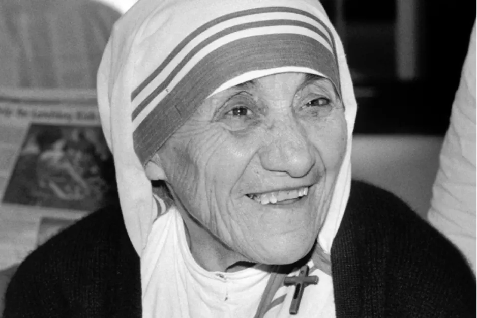 Mother Teresa smiling, wearing her iconic white and blue sari and a cross necklace in a black and white photo.