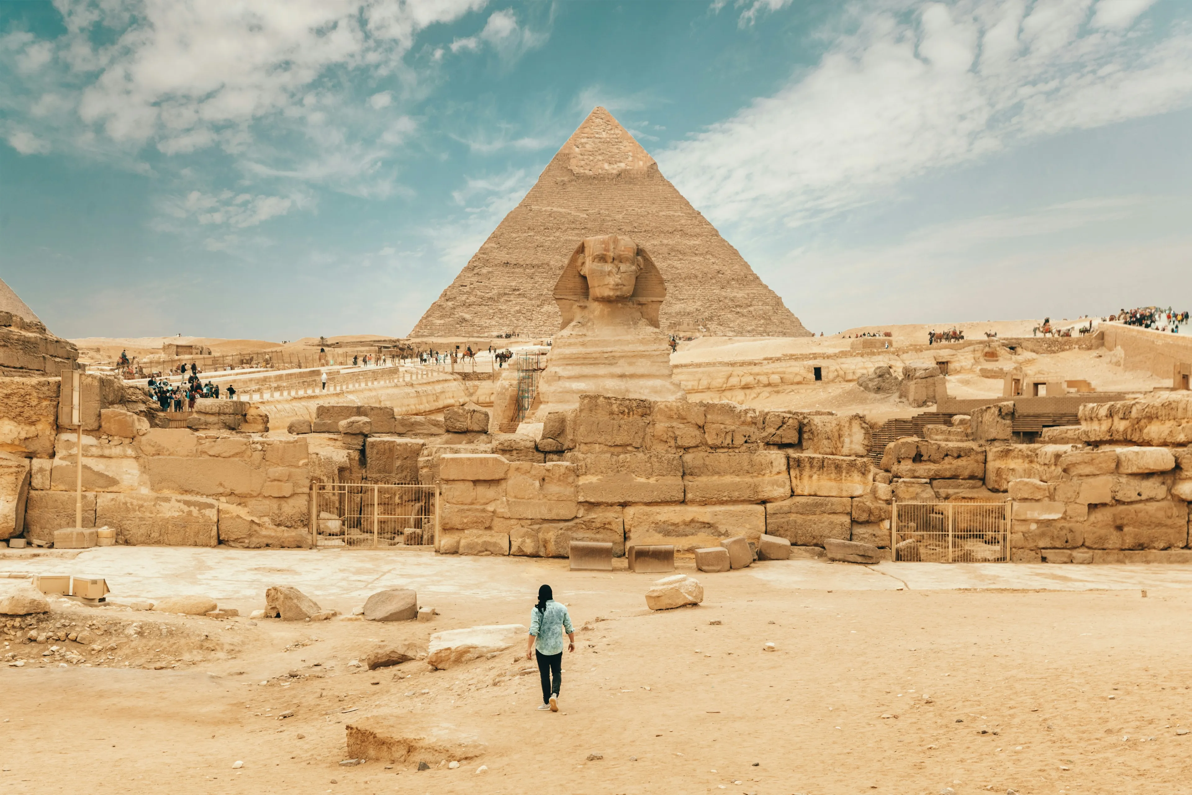 The Great Sphinx and Pyramid of Giza in Cairo, Egypt, with a person walking in the desert under a blue sky.