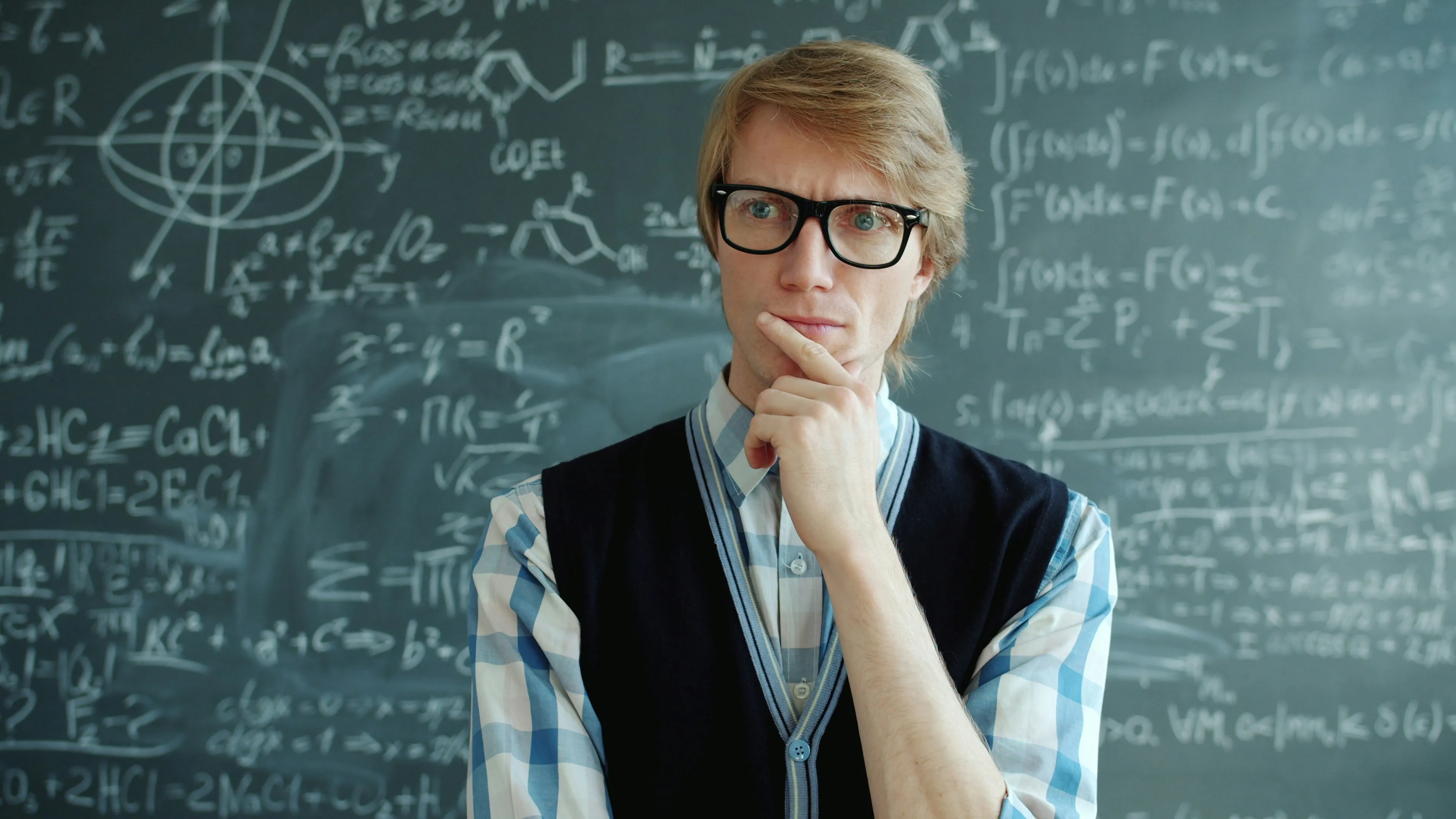 Young man wearing glasses, thinking in front of a chalkboard filled with complex science and math formulas for general knowledge quiz.
