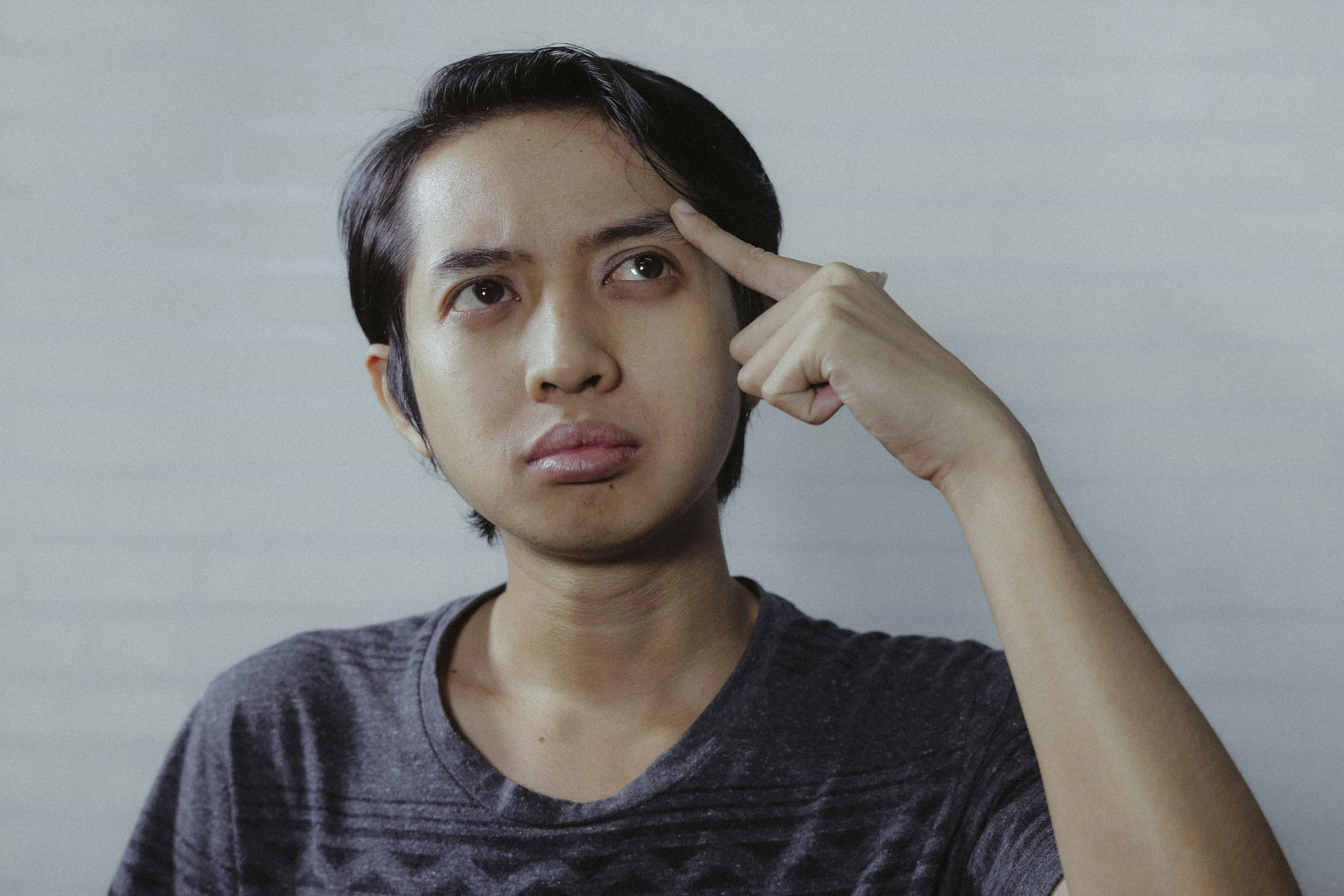 Young man in a gray shirt thinking deeply while pointing to his temple during a general knowledge quiz about the Moon sky color.