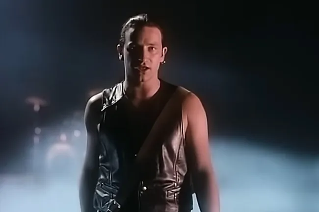 Lead singer of U2 performing With or Without You in a dark smoky setting, wearing a sleeveless leather vest.