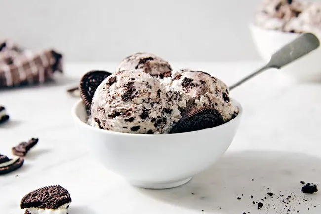 Bowl of cookies and cream ice cream with cookie pieces and a spoon on a white marble surface.