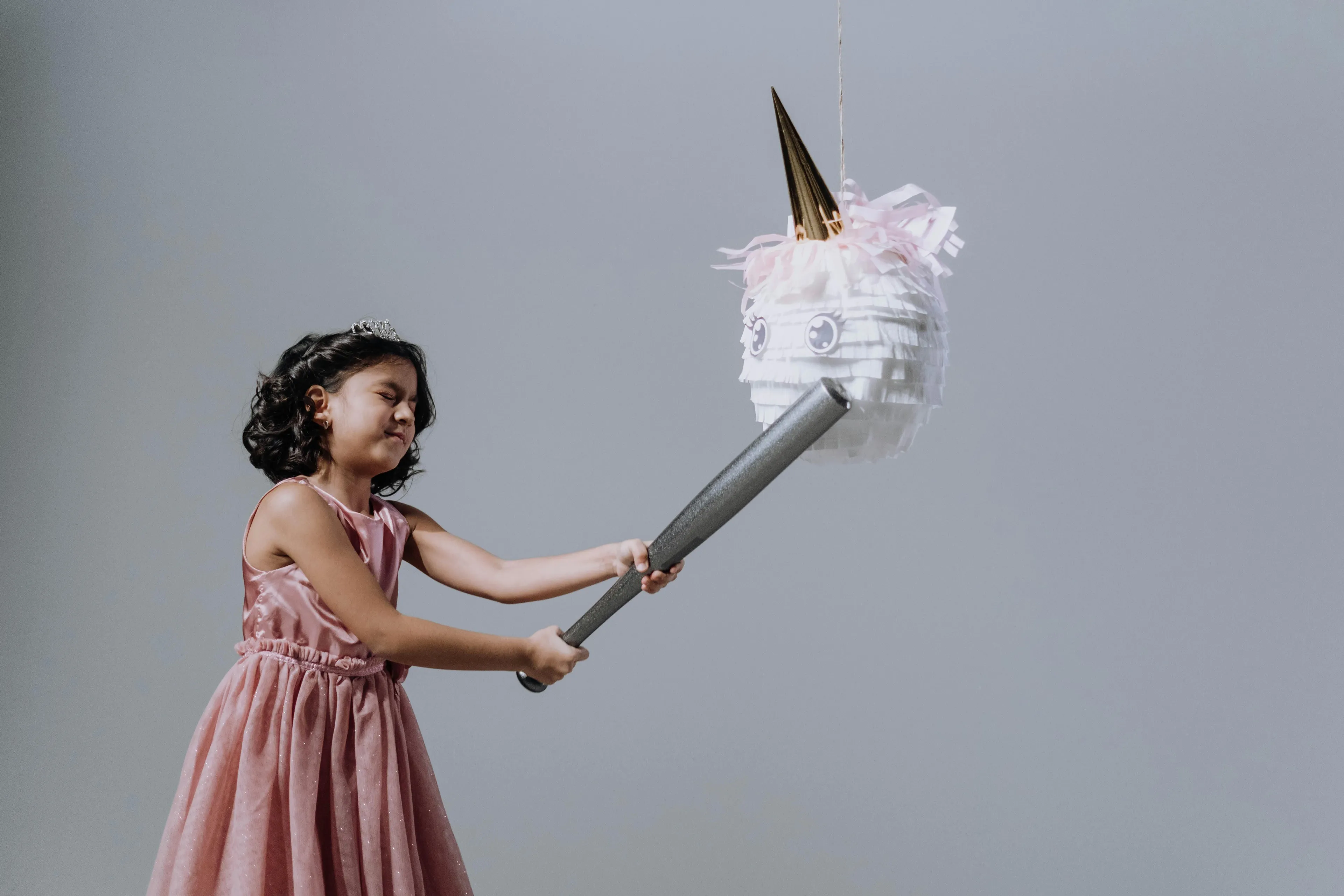 Young girl in a pink dress hitting a unicorn piñata with a bat at a classic kid birthday party activity.