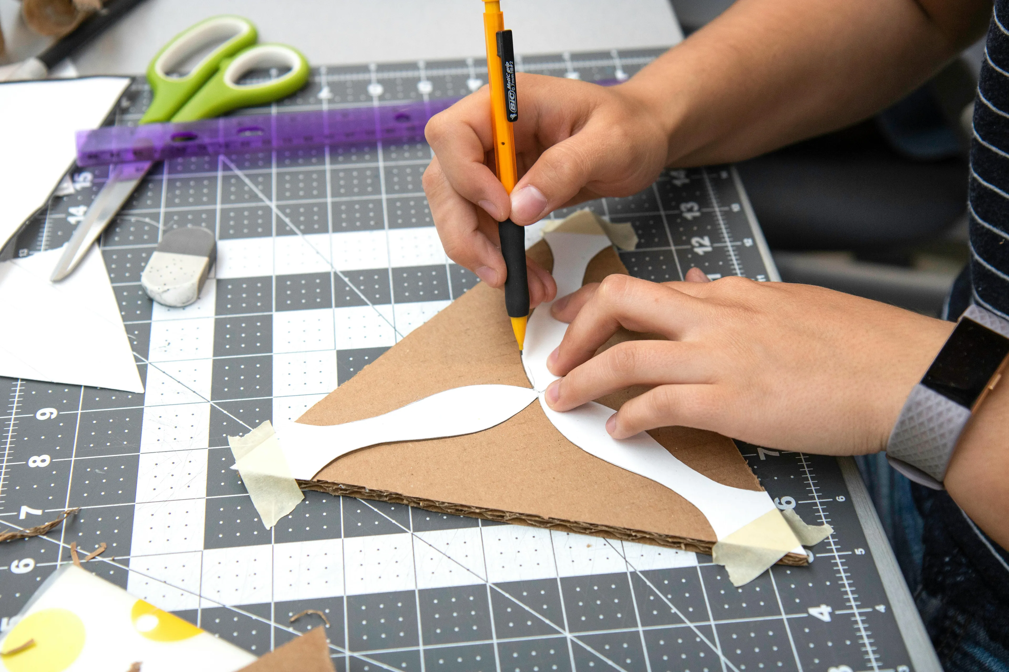 Hands tracing a DIY project template on cardboard using a yellow pencil on a cutting mat with crafting tools nearby