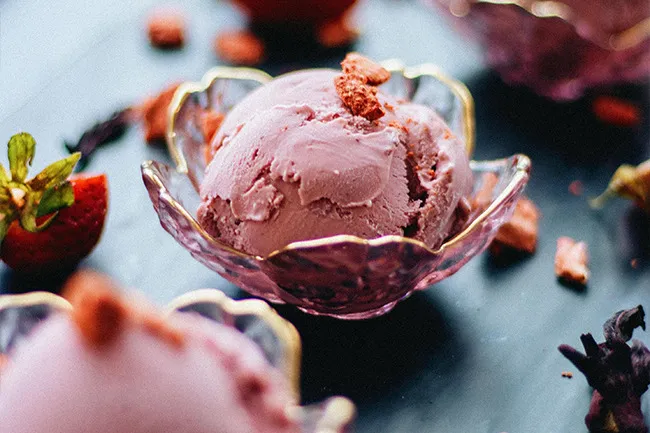 Strawberry ice cream scoop served in a decorative glass bowl with fresh strawberries and chocolate pieces nearby.