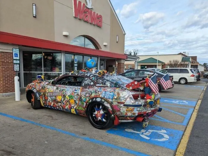 Auto decorado con múltiples stickers y banderas de EEUU estacionado, representación divertida de la vida cotidiana en distintos estados.