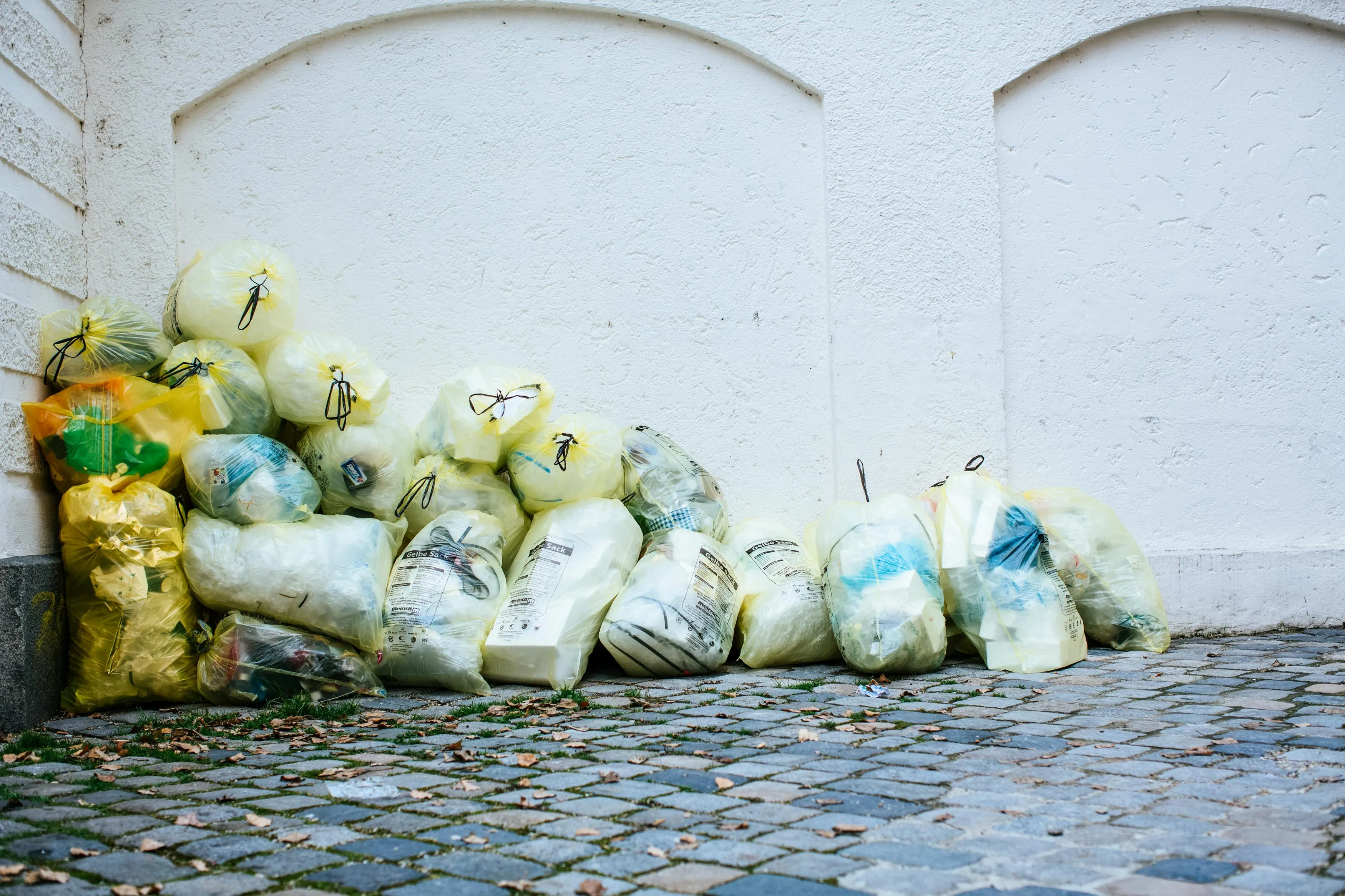Stacks of yellow garbage bags filled with unsold food and packaging lined up against a white wall on a cobblestone street.