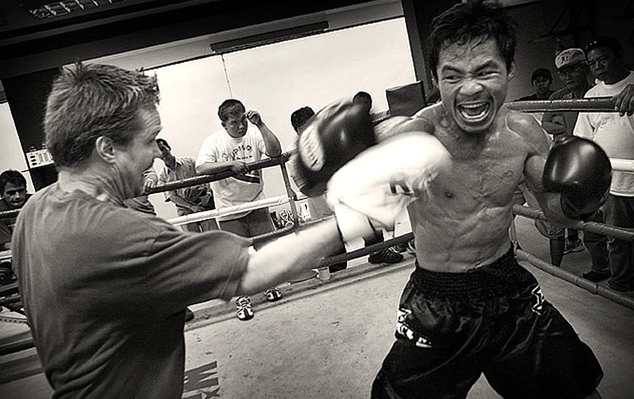 Black and white photo of a boxer training intensely in a gym with a coach hitting focus mitts during a boxing workout.