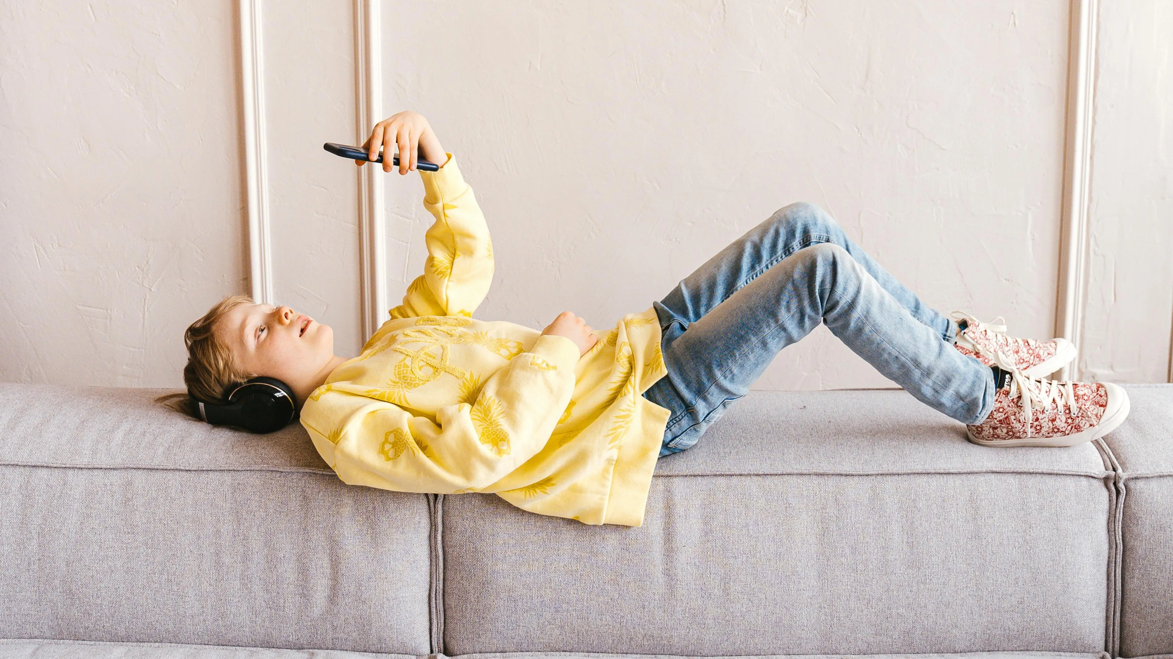 Child wearing headphones and yellow sweatshirt lying on a couch using smartphone related to countries that were first quiz.