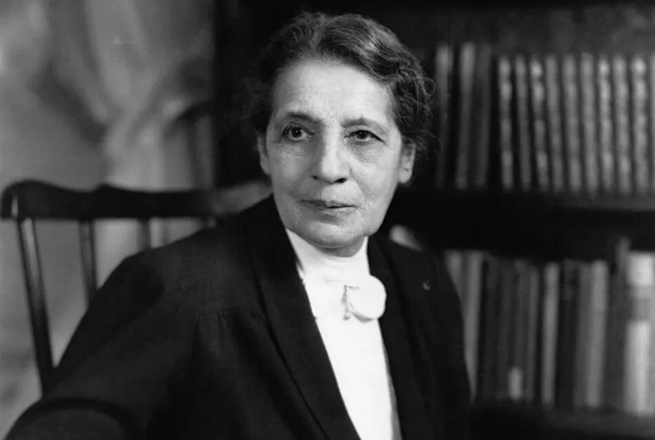 Black and white portrait of a woman in formal attire seated in front of a bookshelf, representing Nobel Prize history.